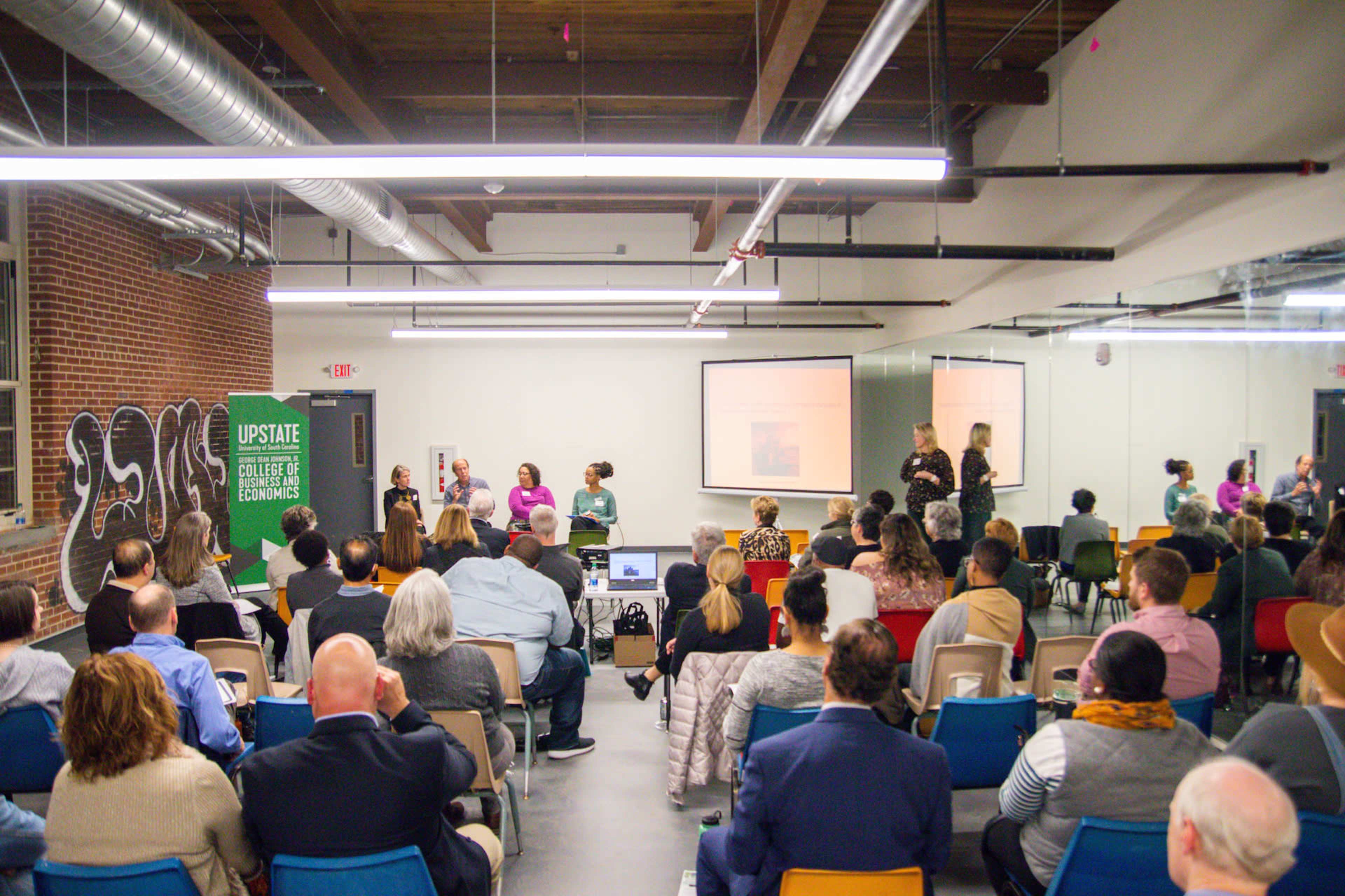 A diverse group of people attends a presentation in a spacious room with brick walls and modern lighting.