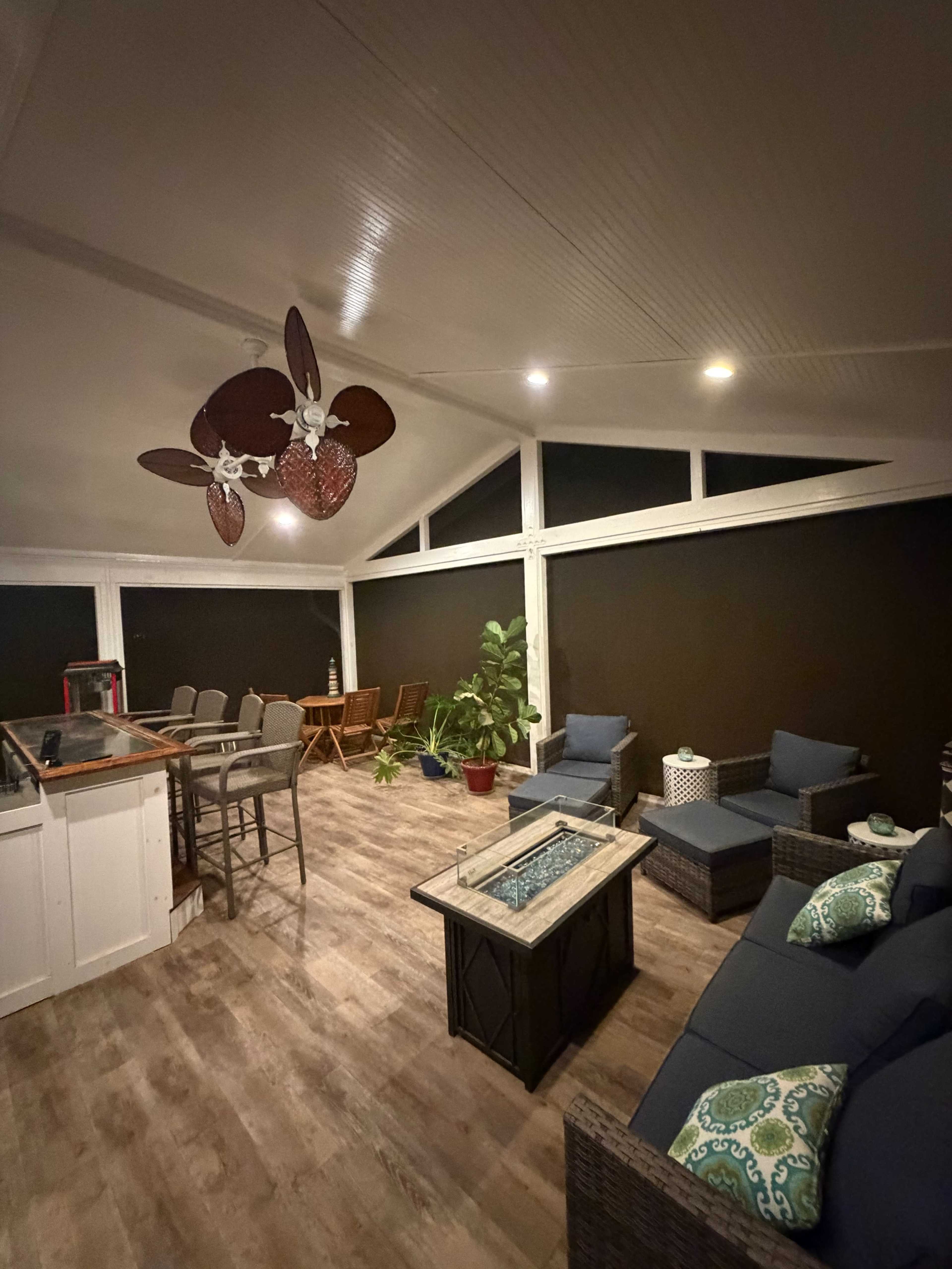 The image shows a modern screened patio with seating arrangements, a dining table, and a fire pit, all illuminated by overhead lights.