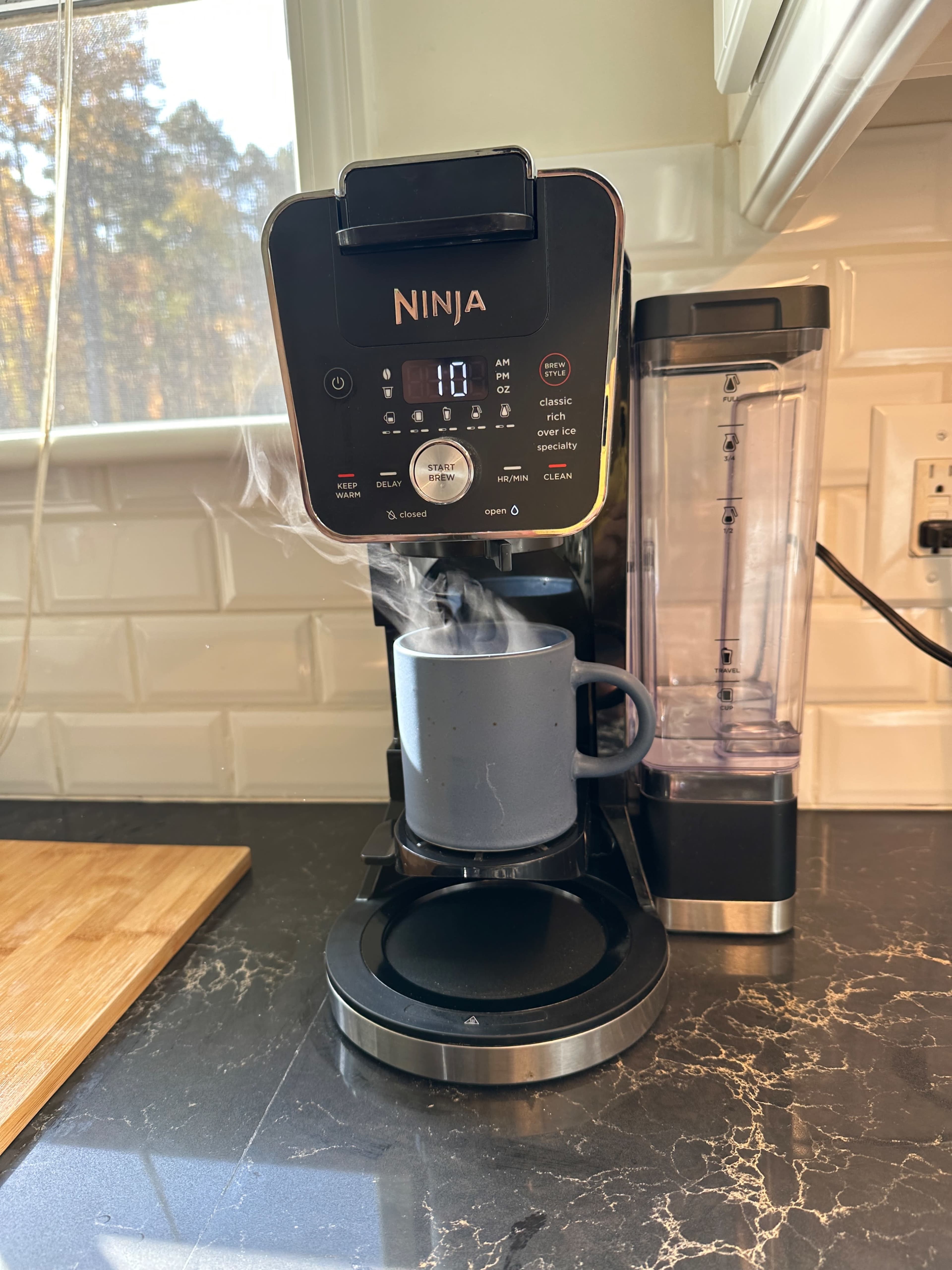 A coffee maker with a steaming mug is positioned on a kitchen countertop next to a water reservoir.