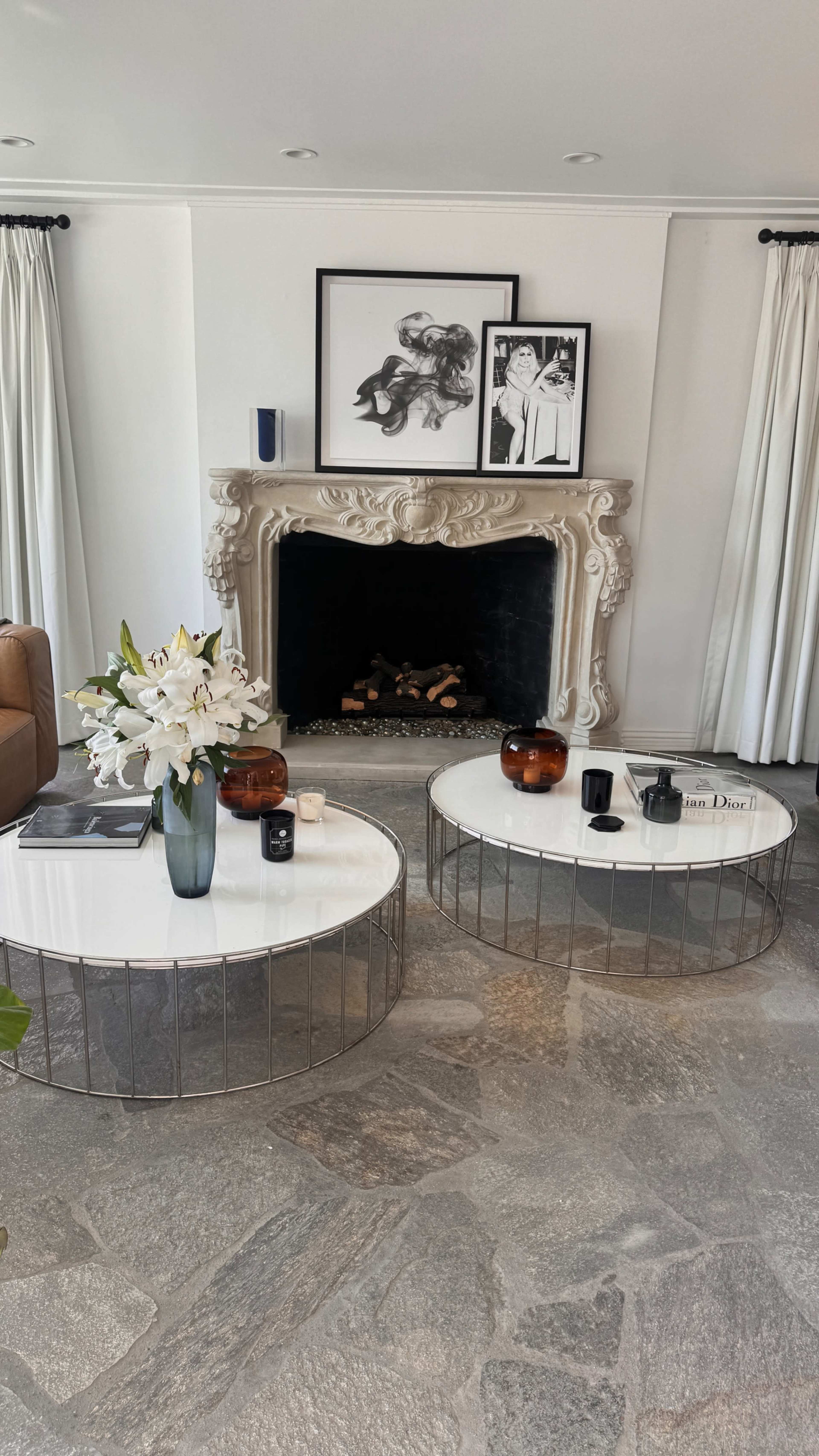 The image shows a stylish living room featuring two round coffee tables, a decorative fireplace with white stone detailing, and large windows dressed with white curtains.