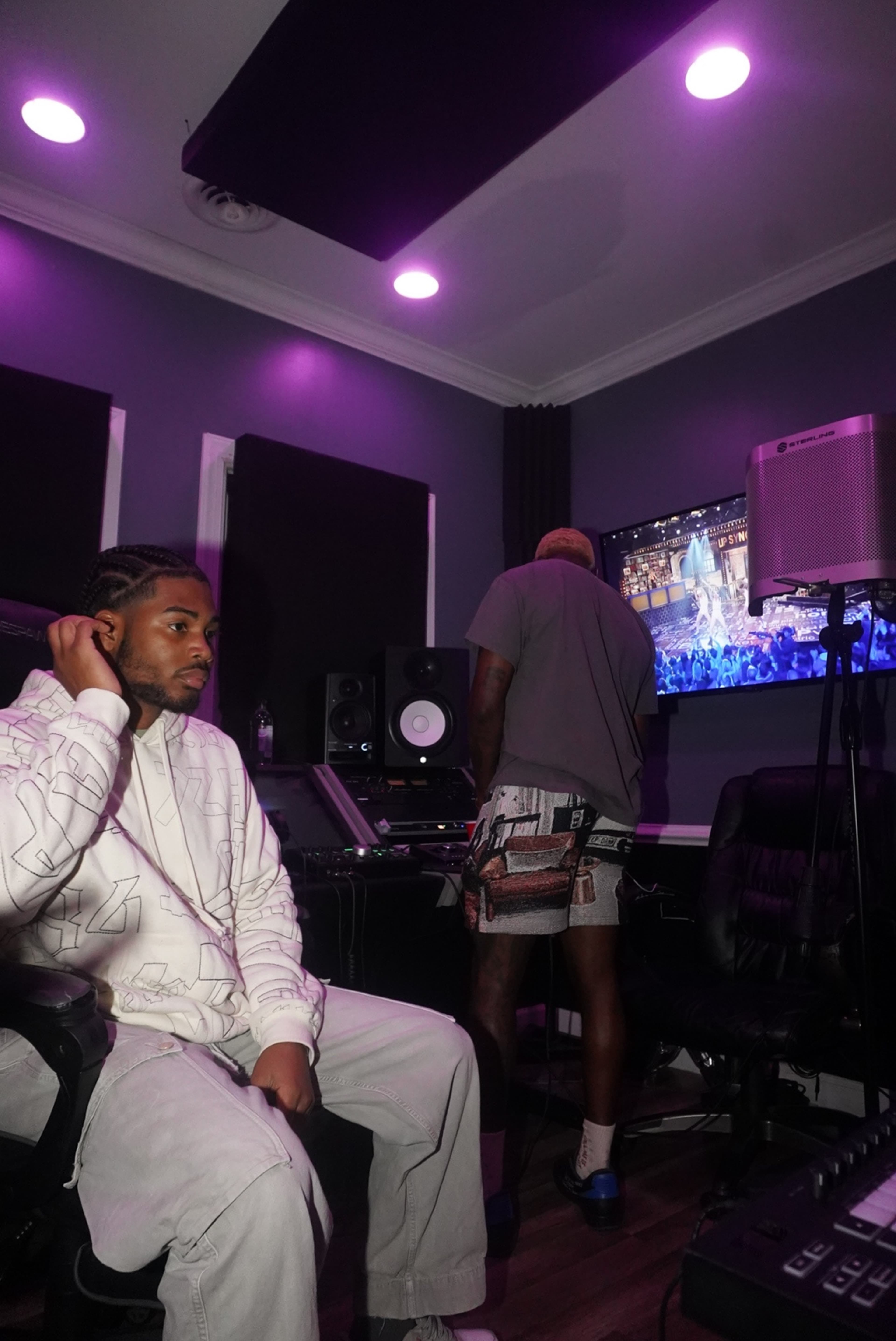 Two individuals are in a recording studio, one sitting in a chair while the other stands near equipment, with a screen displaying a performance in the background.