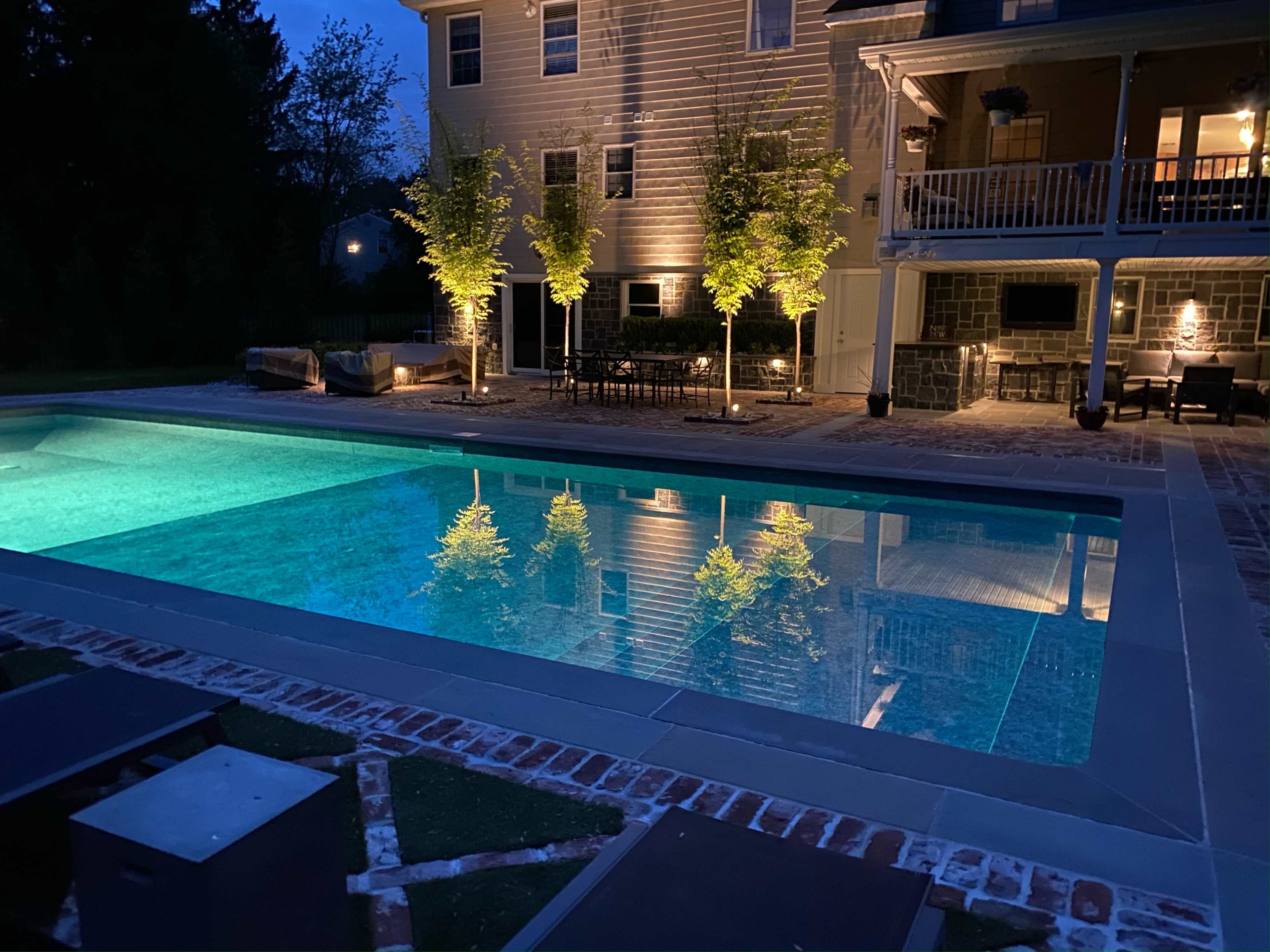 A well-lit backyard pool area with a rectangular pool, surrounded by chairs and a dining table, against a backdrop of a two-story house at dusk.
