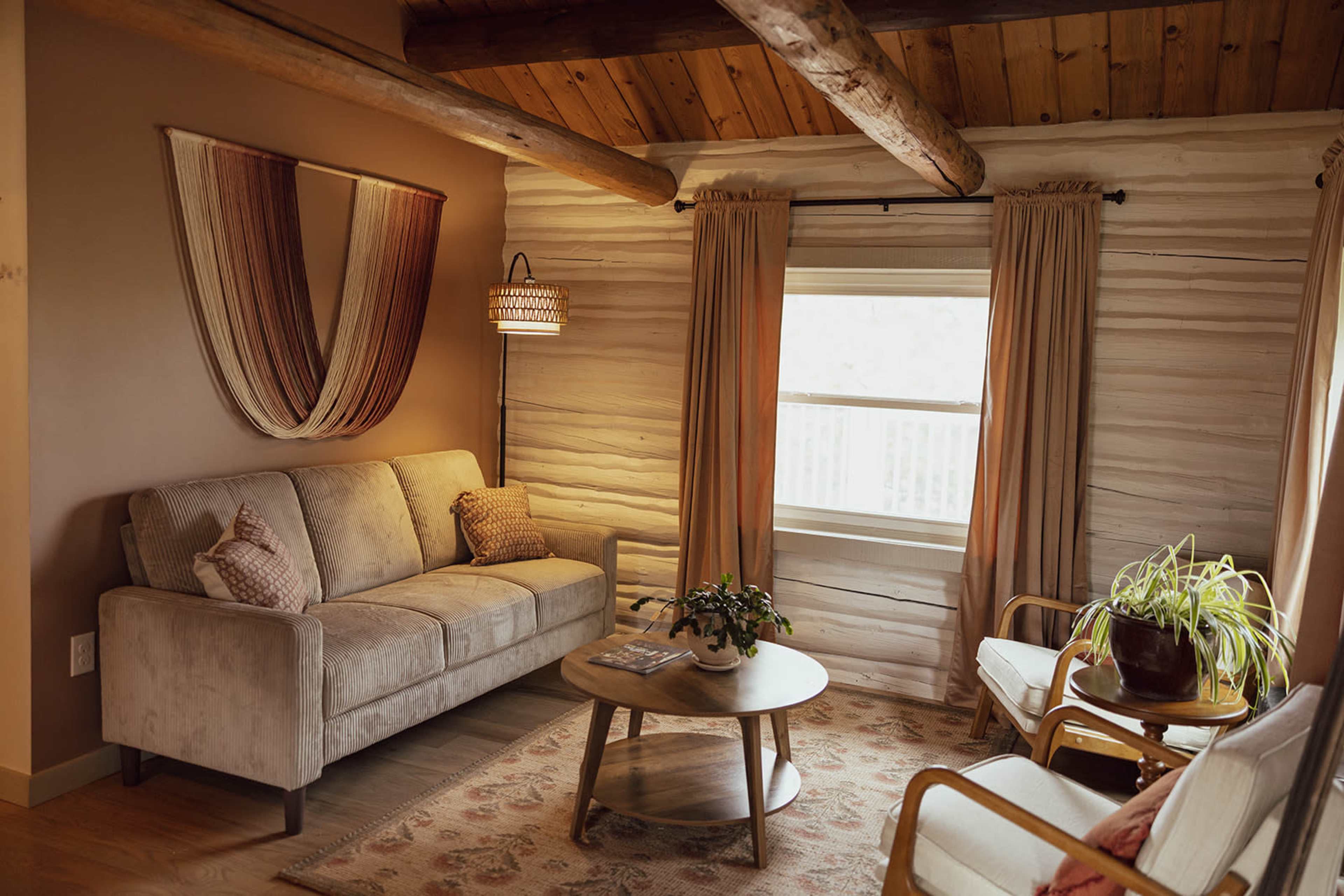 The image shows a cozy living room with a beige couch, wooden beams on the ceiling, a round coffee table, and a plant near a window.