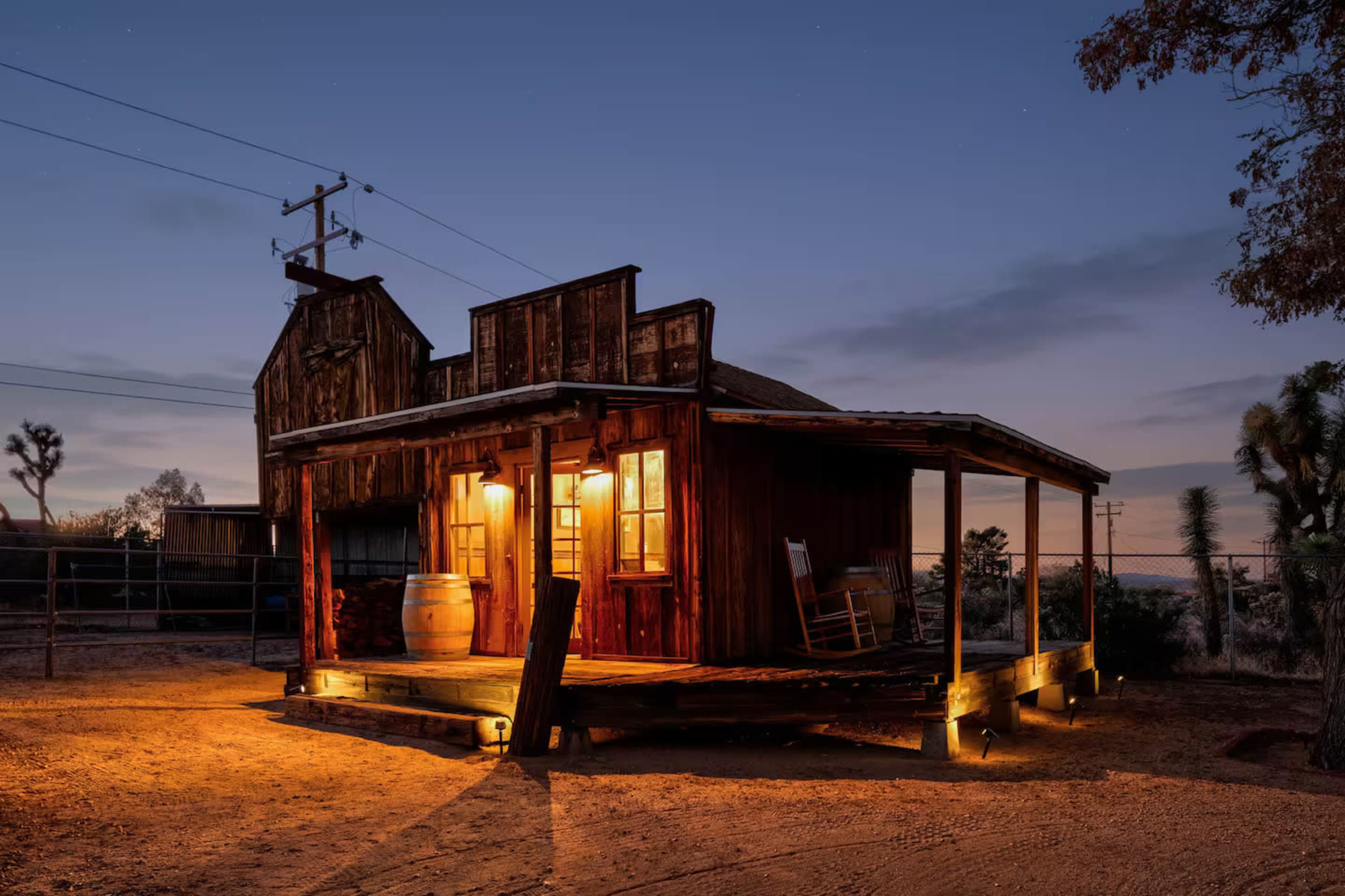 A rustic wooden cabin is illuminated at dusk, featuring a porch with a rocking chair and a barrel set against a desert landscape.