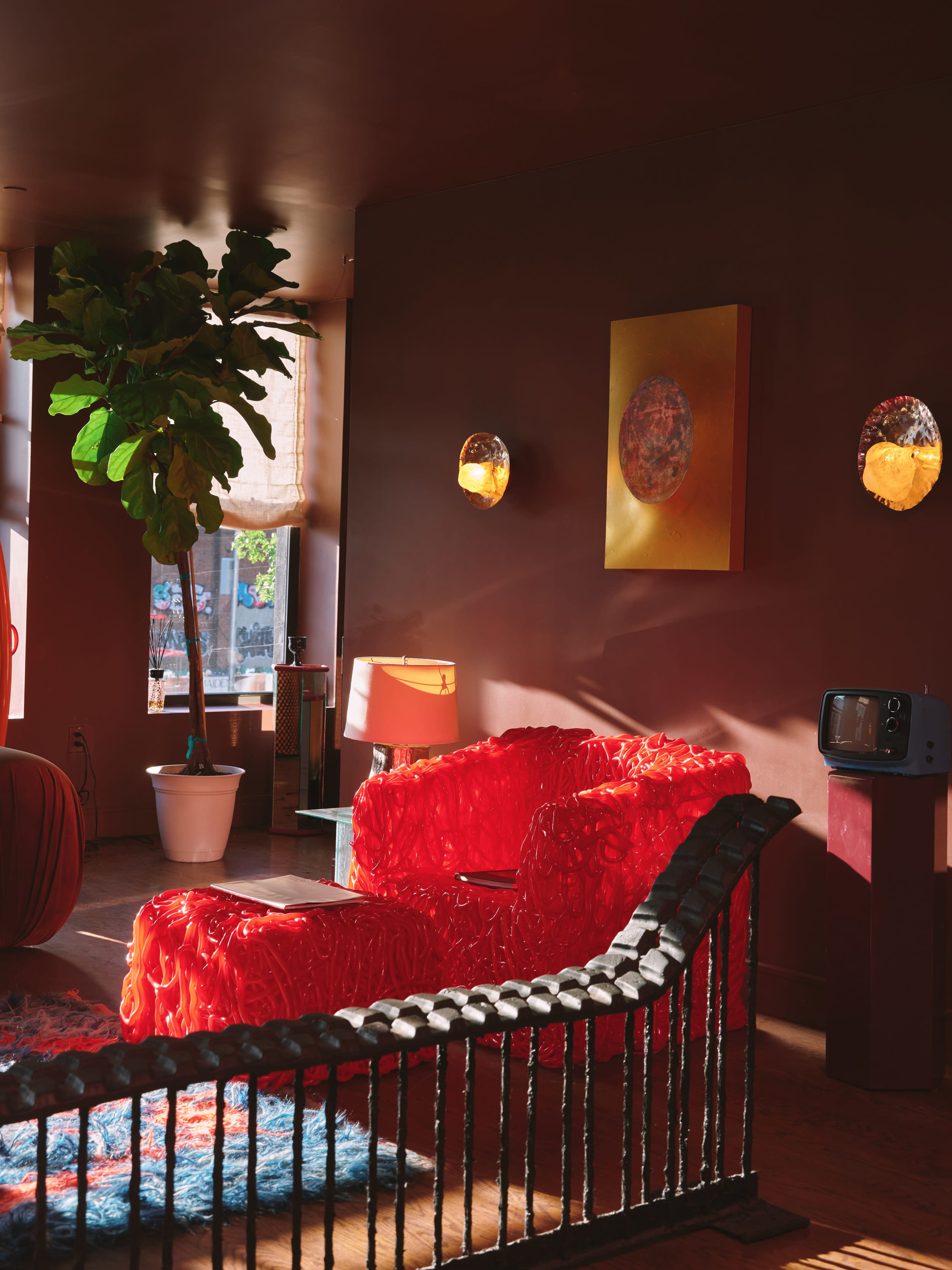 The image shows a cozy living room featuring bright red furniture, a large potted plant, and warm lighting against a brown wall.