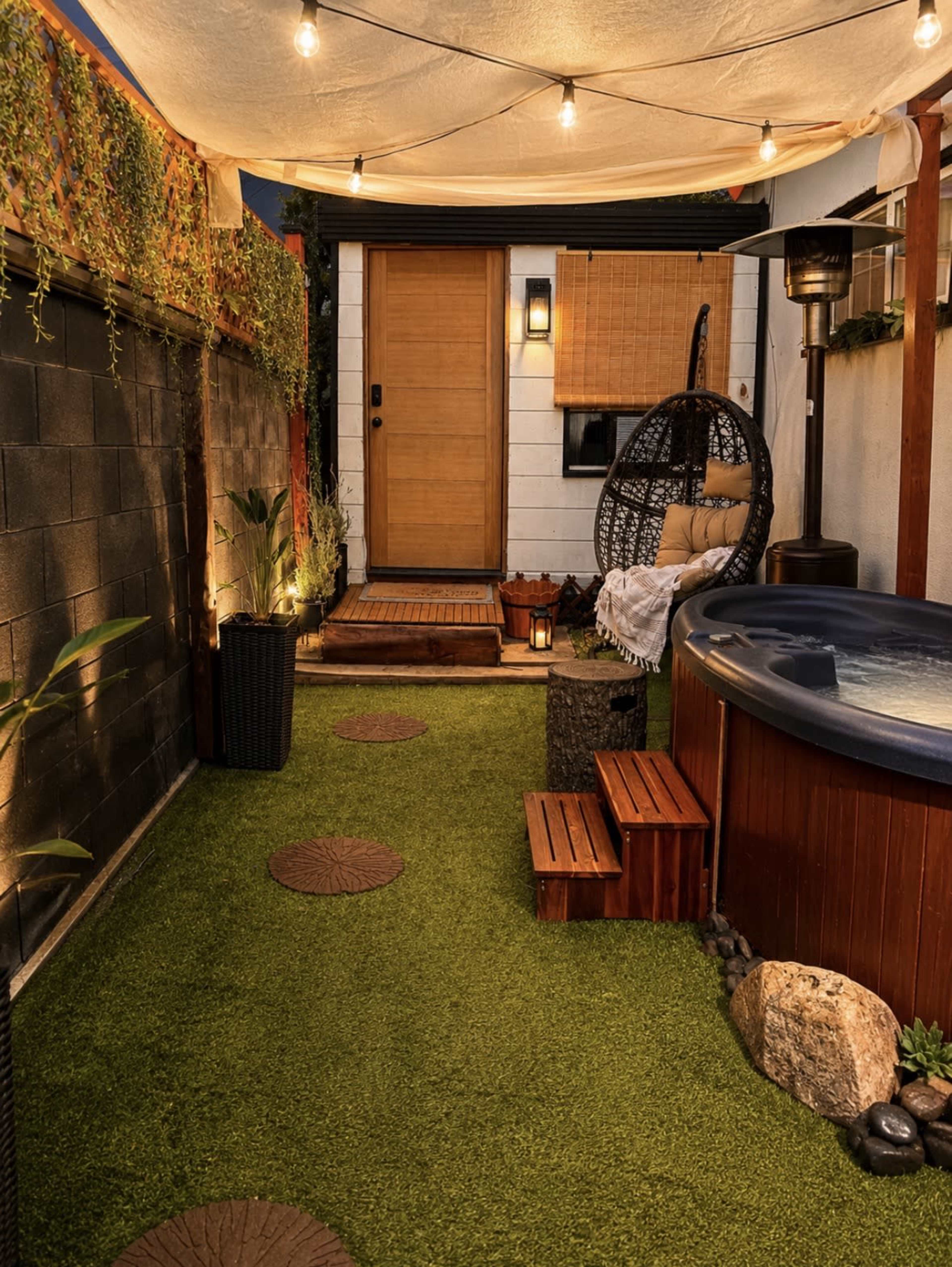 The image shows a landscaped backyard area featuring a hot tub, decorative stone steps, and a wooden door, all illuminated by soft outdoor lighting.