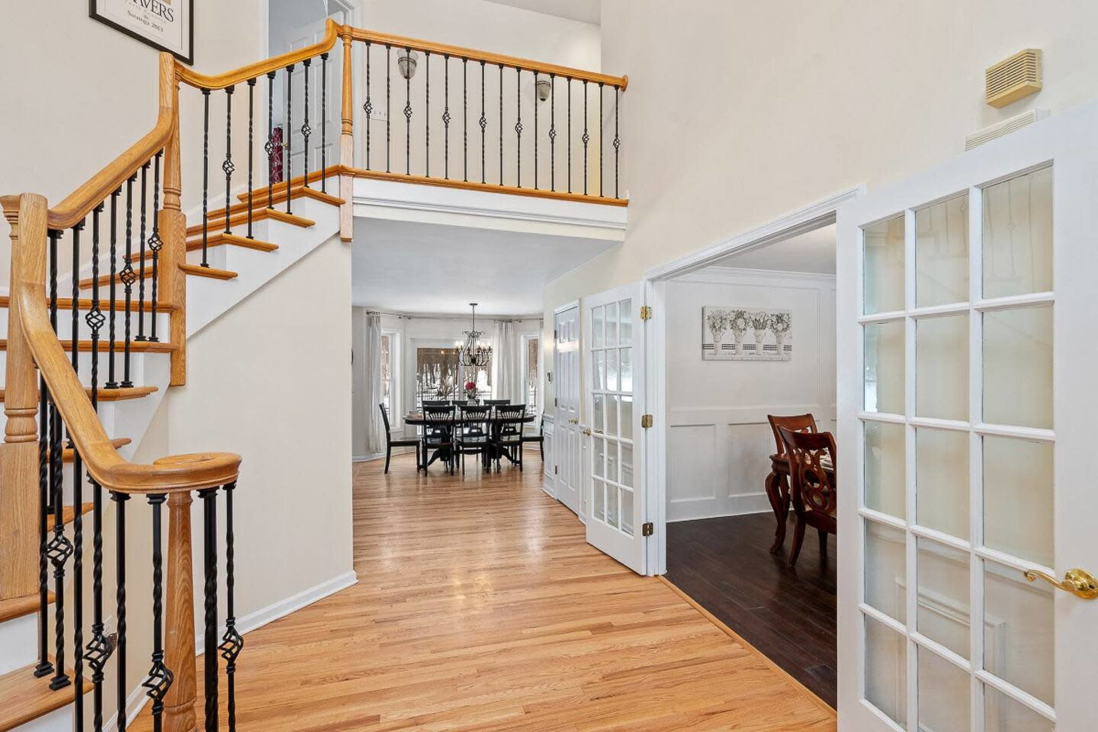 A foyer features a wooden staircase with black railings, leading to an upper level, and opens to a dining area visible through glass-paneled doors.
