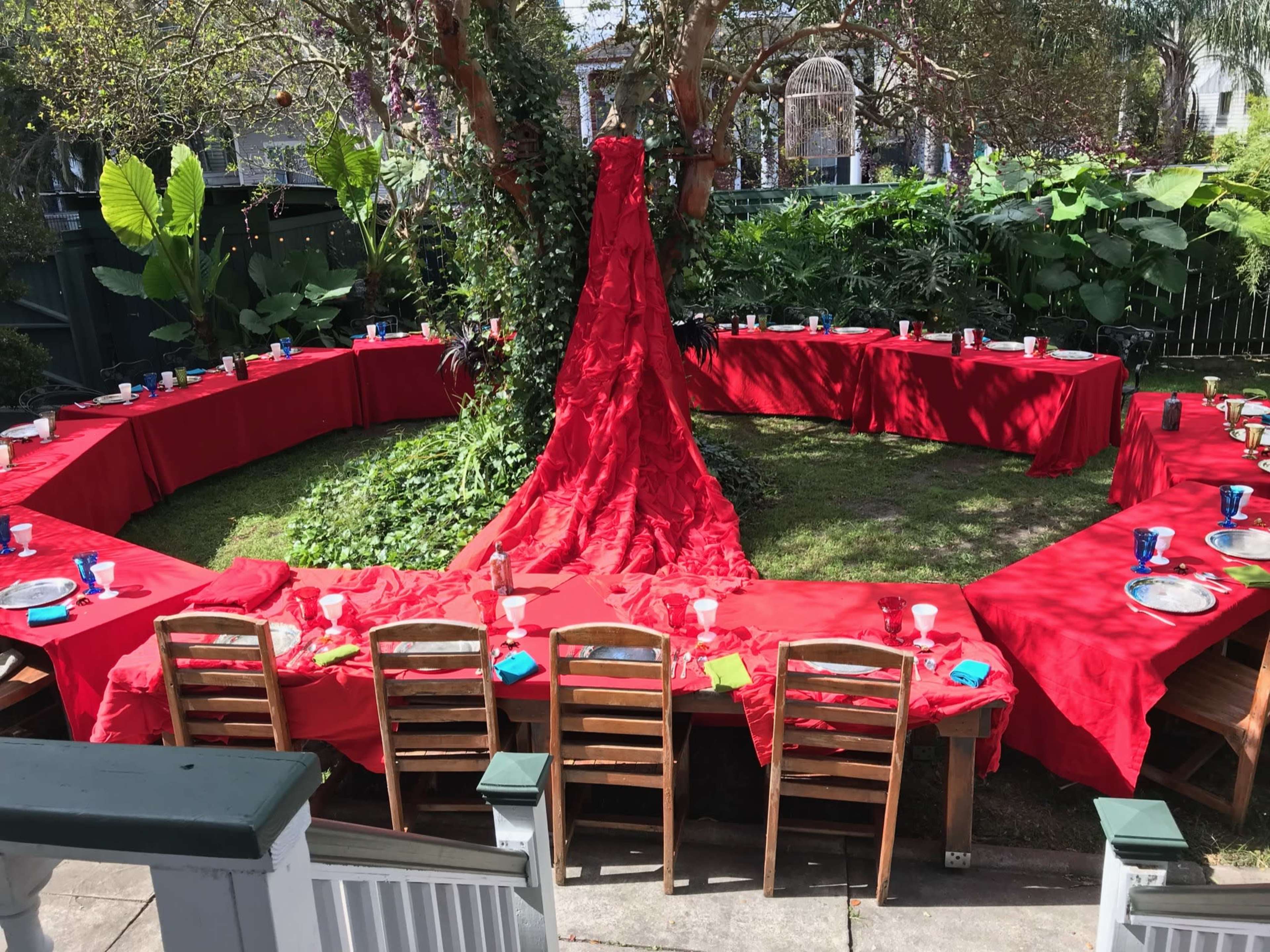 A circular arrangement of tables covered with red tablecloths is set up under a tree in a garden, surrounded by lush greenery.