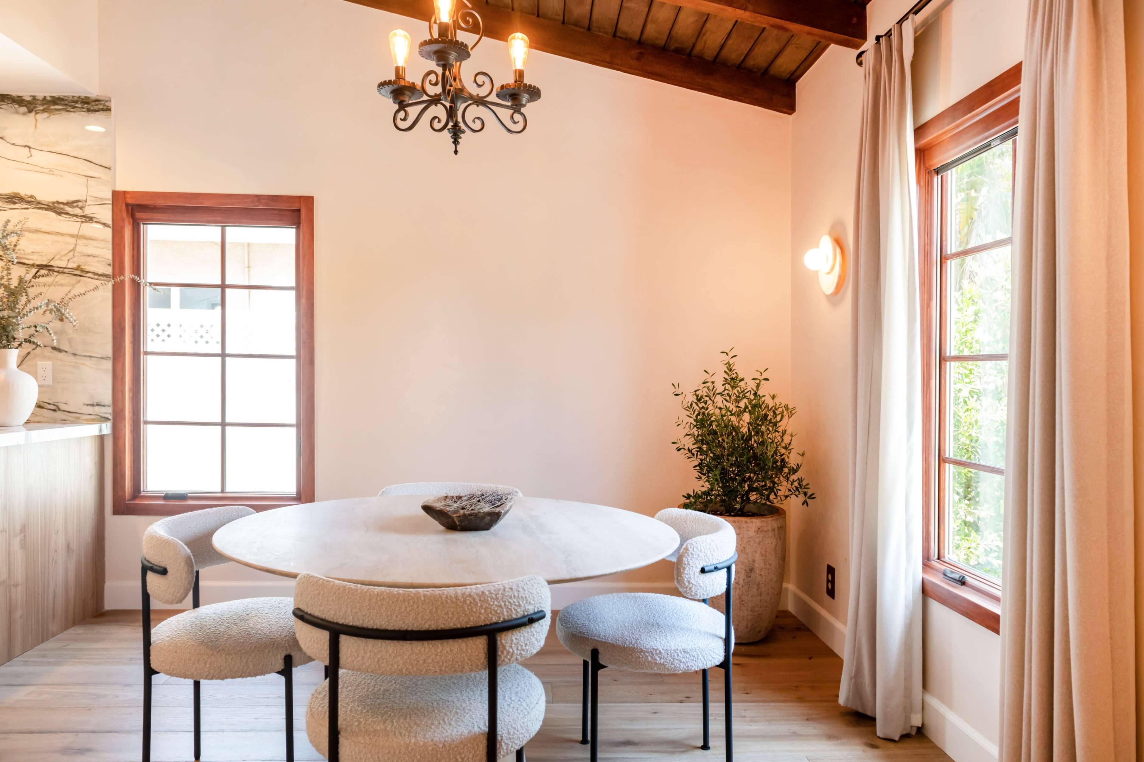 A round dining table with four upholstered chairs is positioned near a window in a light-colored room with wooden accents and a chandelier.