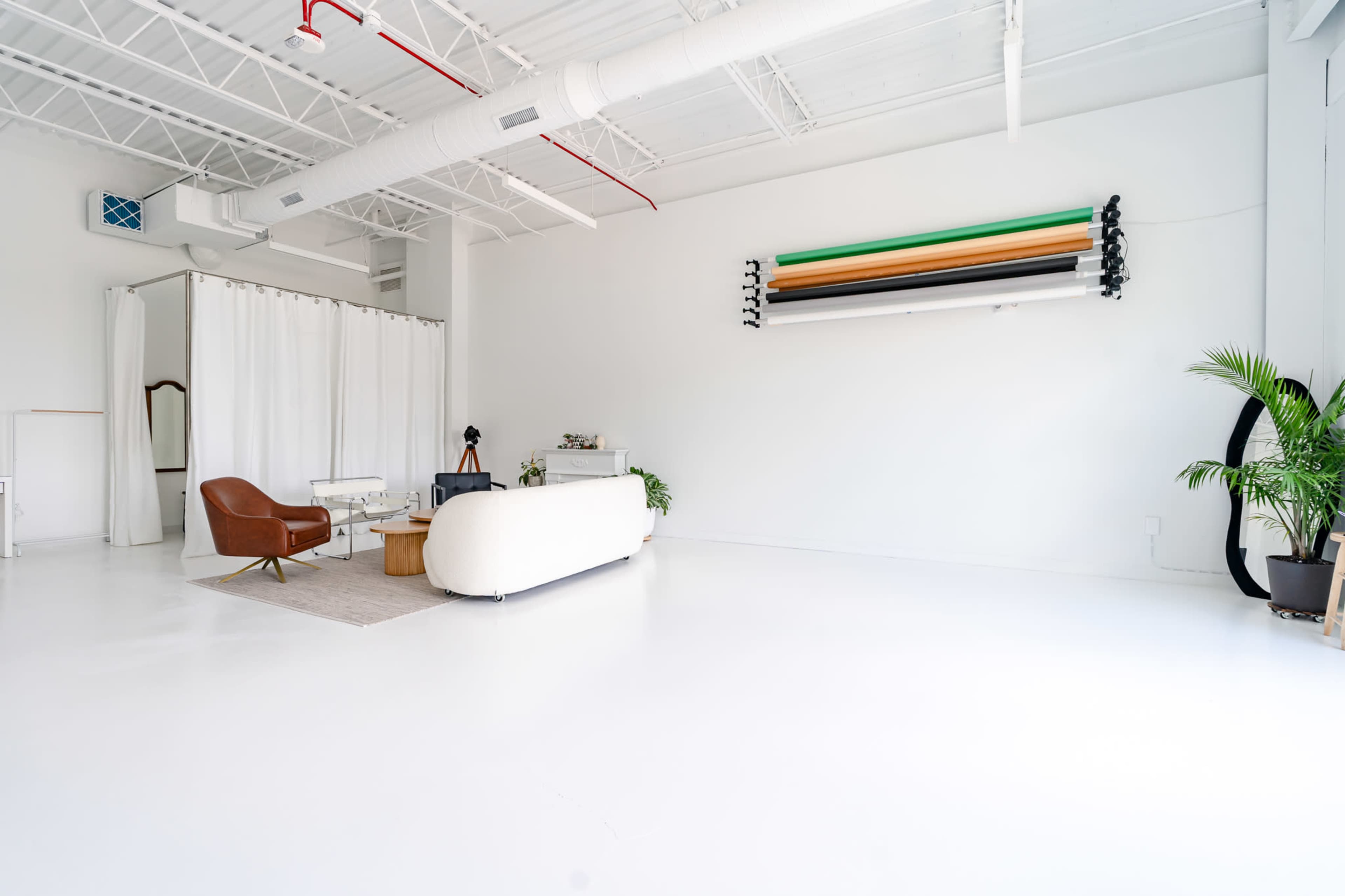 The image shows a spacious, brightly lit studio with white walls and flooring, featuring a cream-colored sofa, a brown armchair, a coffee table, and a decorative plant.