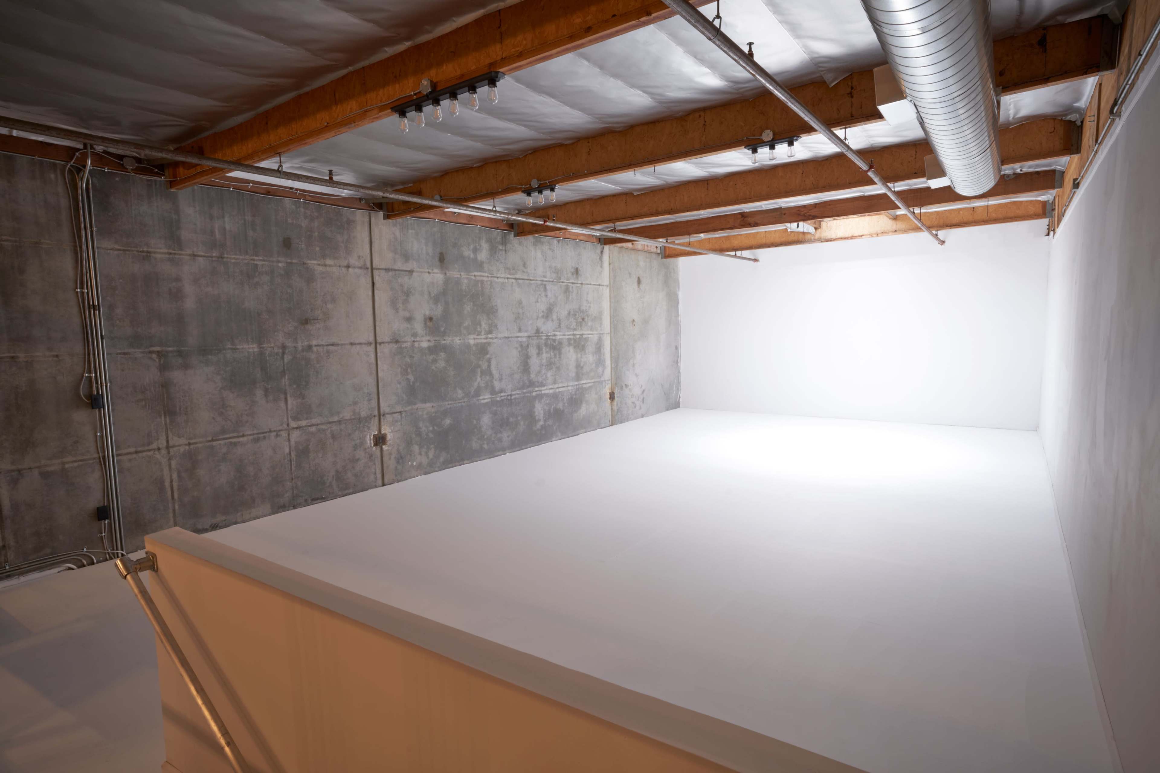 A spacious, empty studio with wooden beams and concrete walls, featuring a bright, white corner illuminated by soft light.