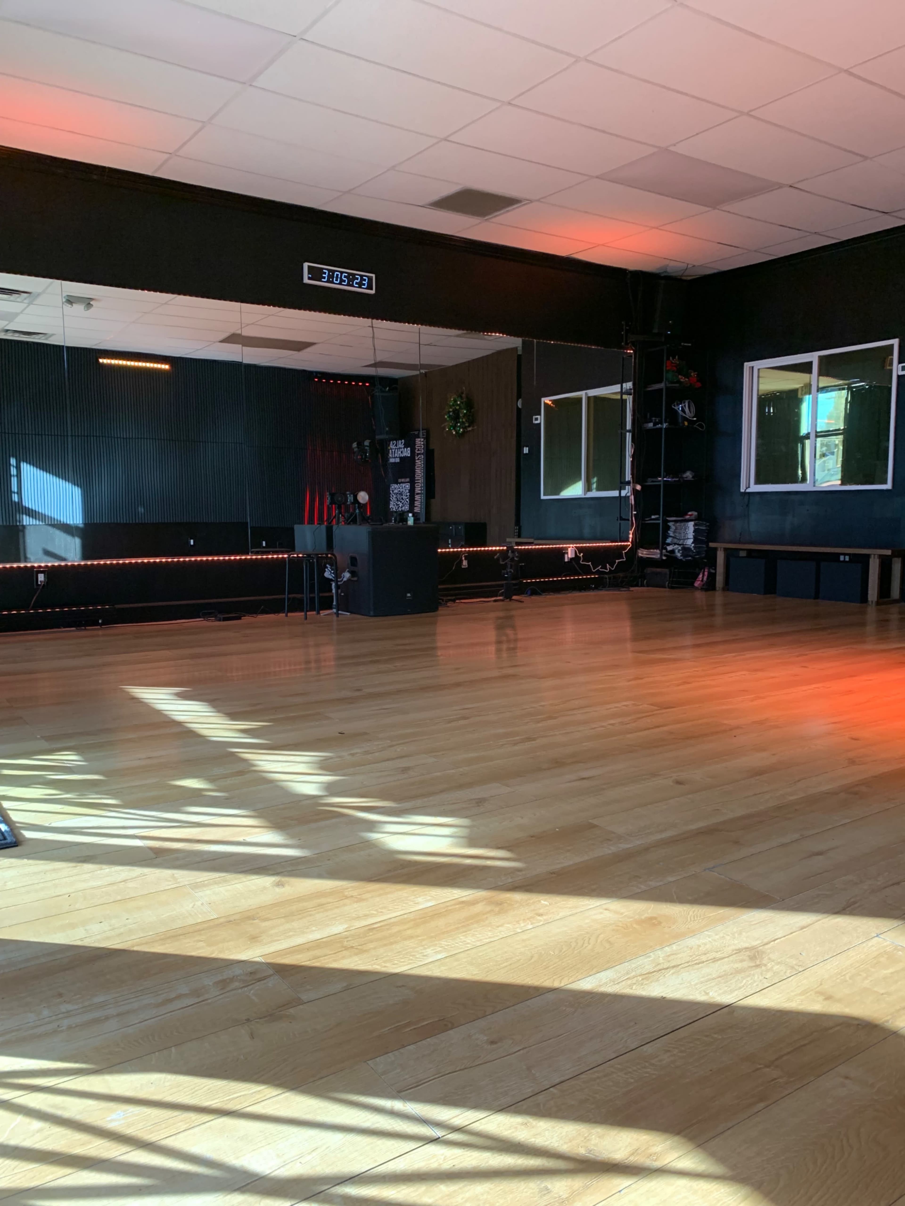 The image shows an empty fitness studio with wooden flooring, a large mirror, and windows illuminated by sunlight.