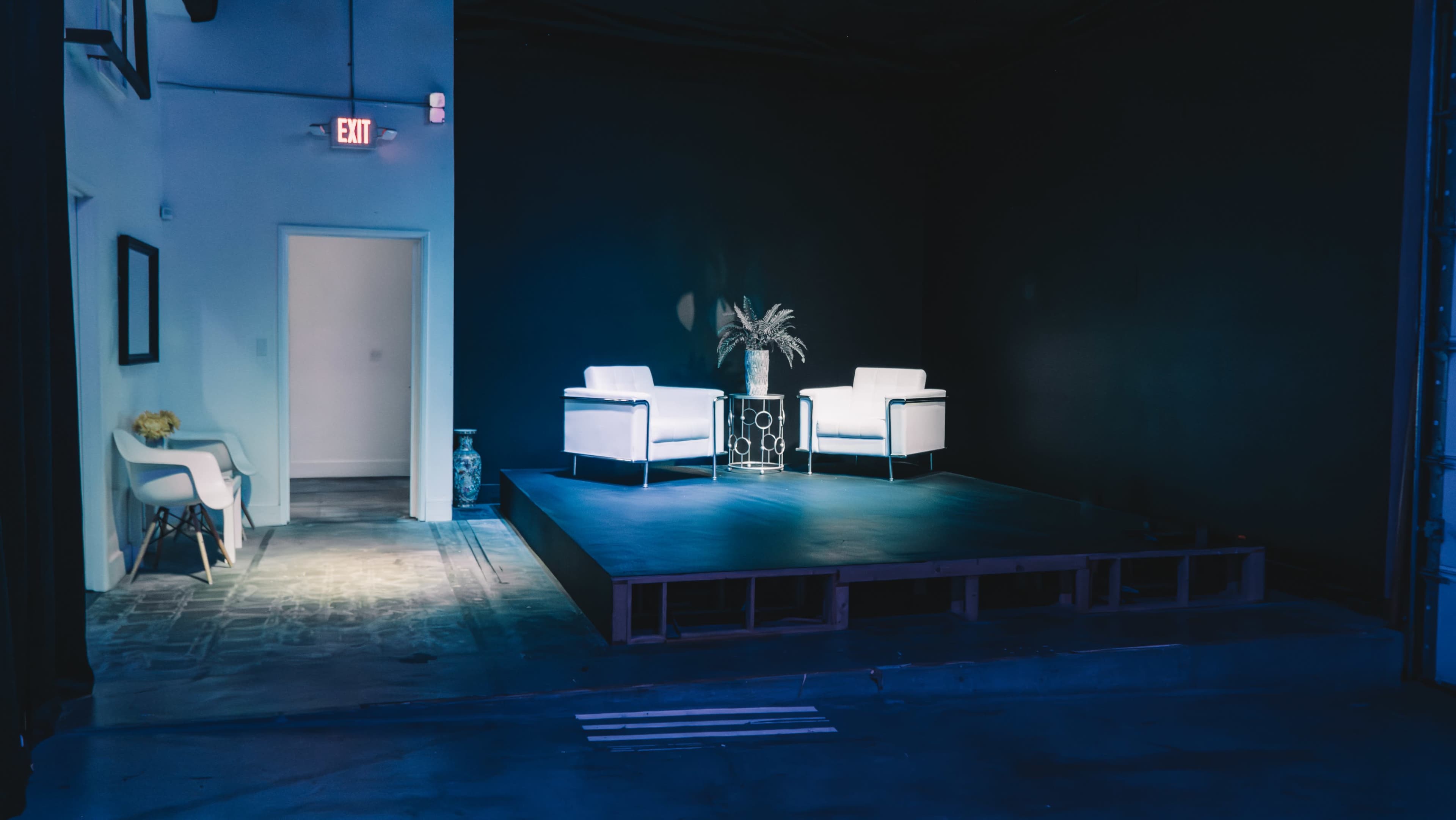 A minimalist stage setup features two white chairs and a plant on a small table, illuminated in a dark room with a doorway on the left.