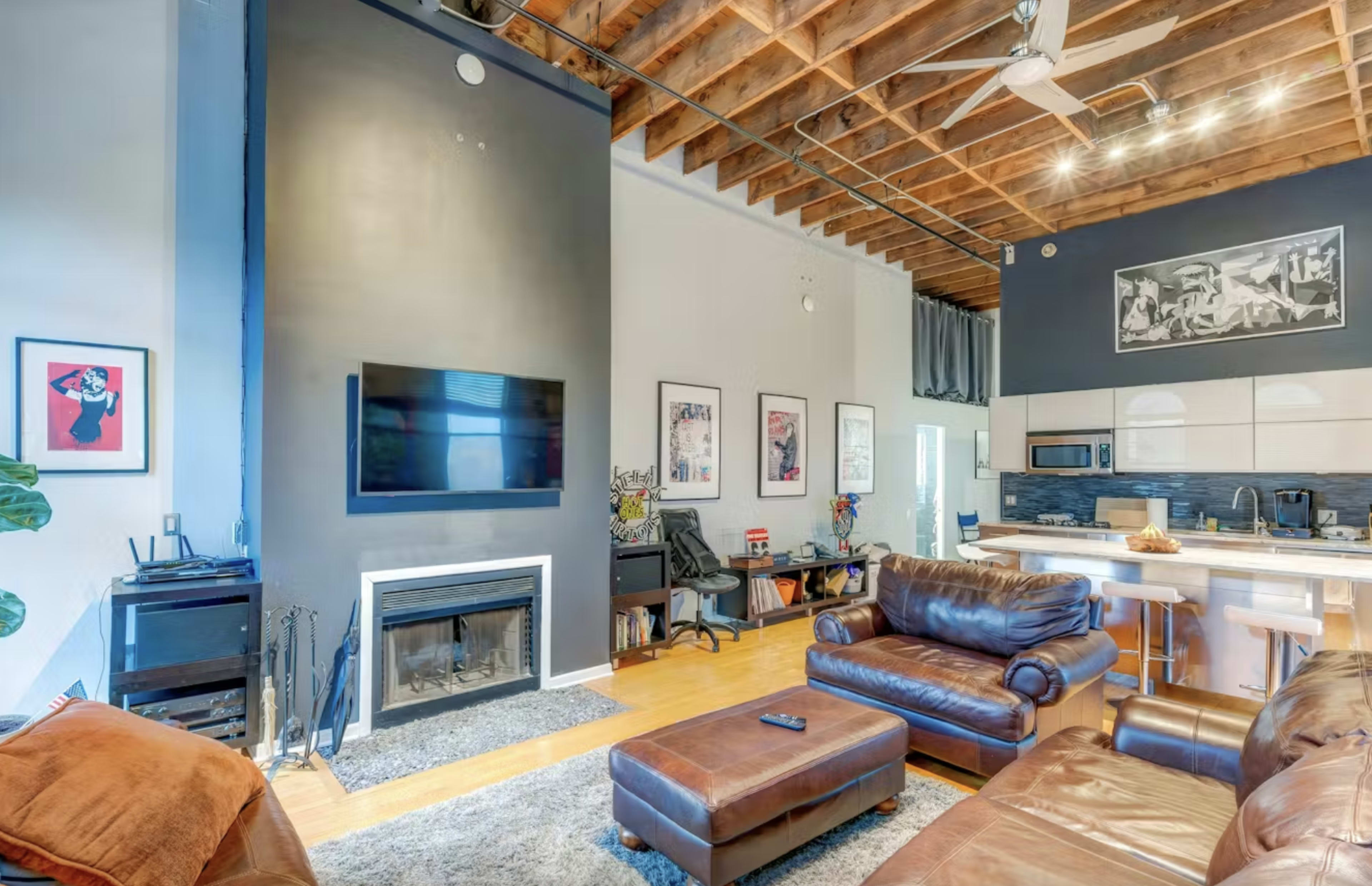 The image shows a modern living room featuring a seating area with leather sofas, a wall-mounted TV, and a fireplace, set against a backdrop of exposed wooden beams and artistic wall decor.