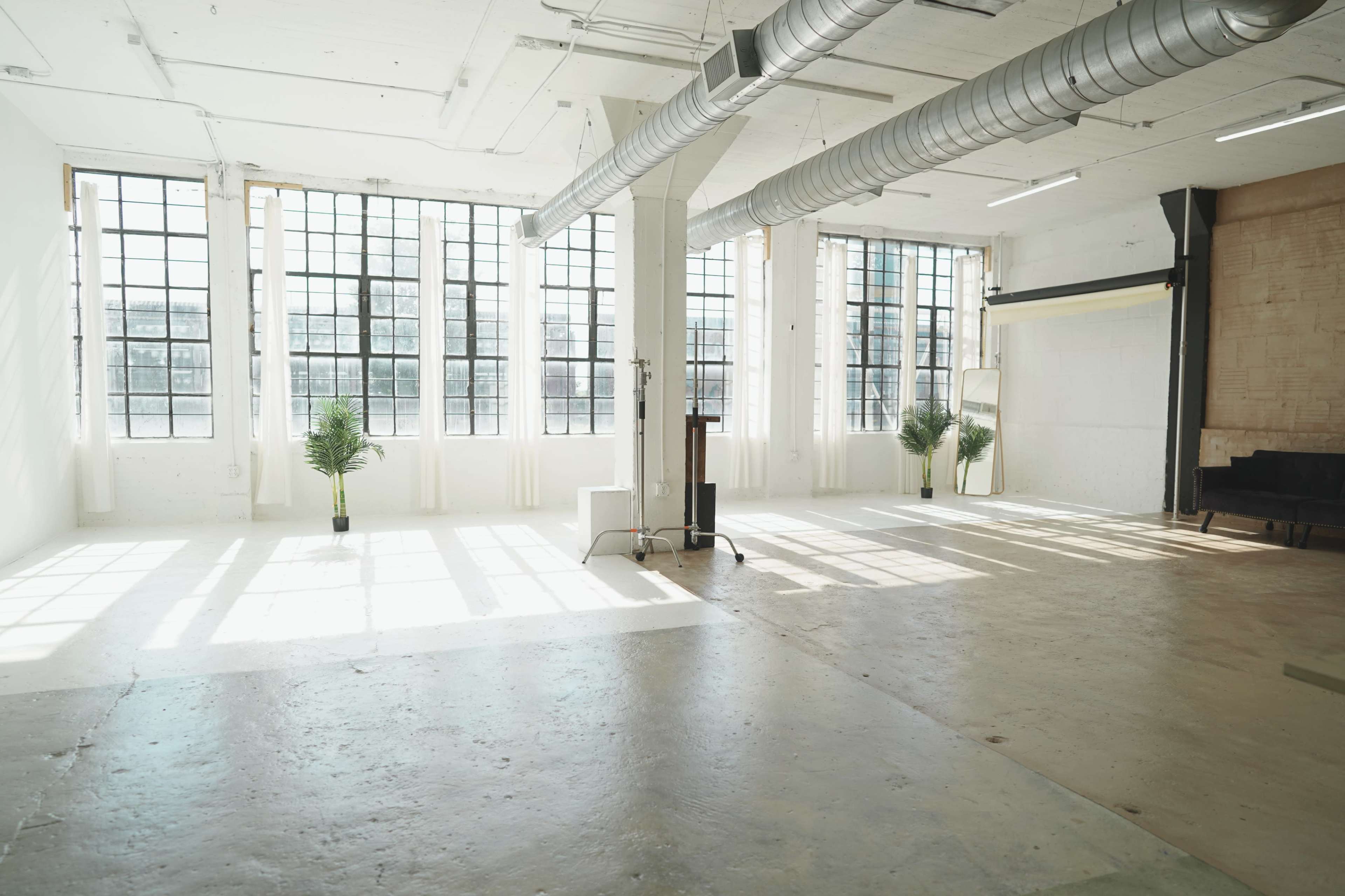 The image shows a spacious, well-lit room with large windows, a concrete floor, and a few potted plants.