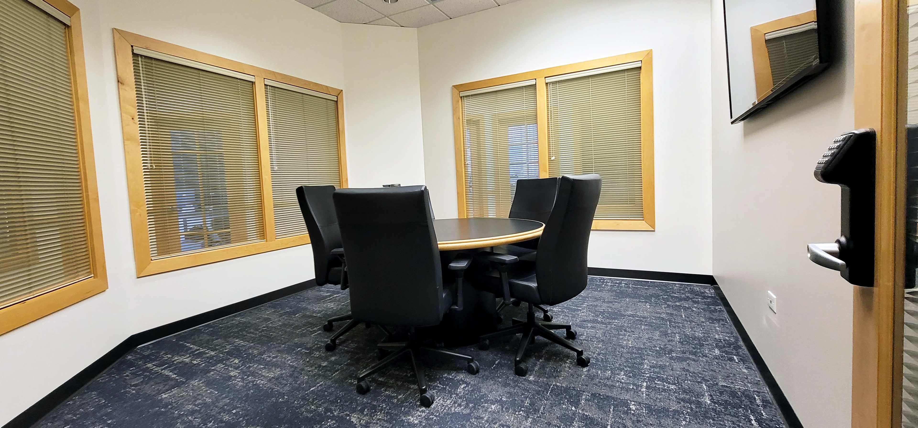 A small conference room features a round table surrounded by four black chairs and windows with blinds.