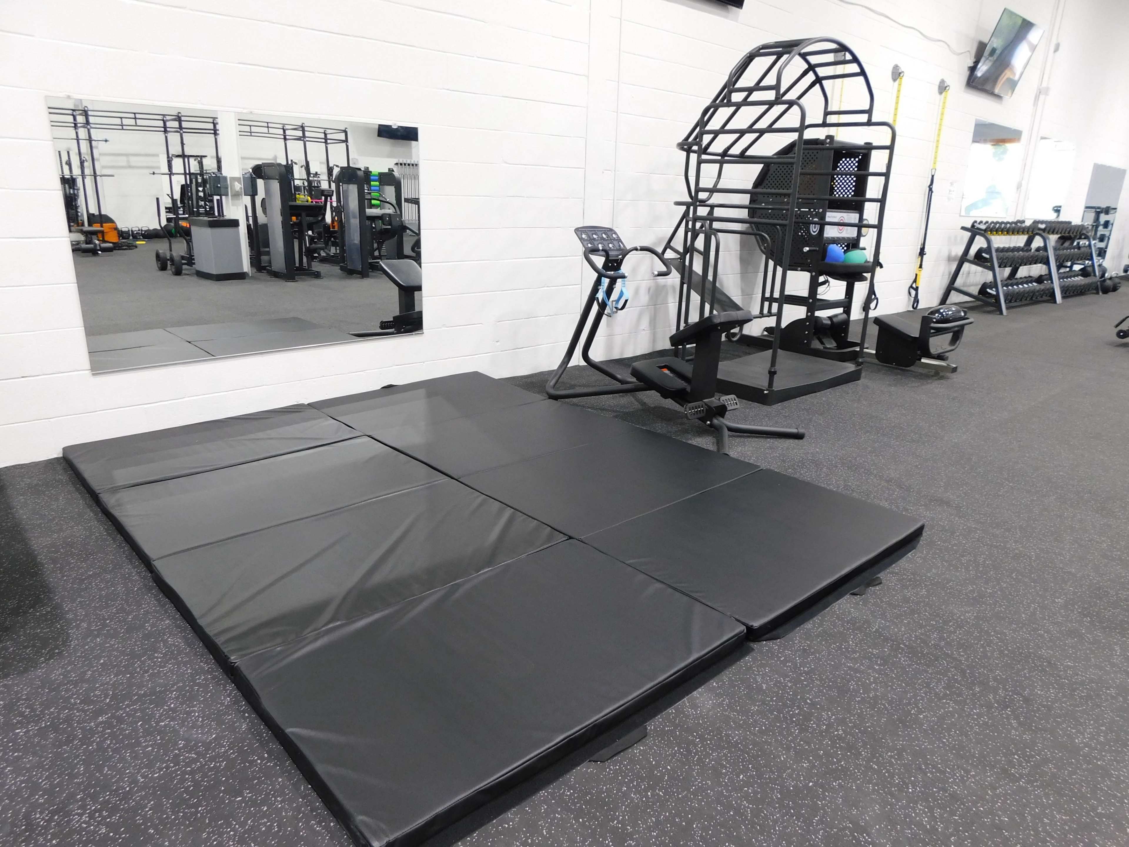 A gym interior features a black exercise mat next to a multi-functional workout machine and a large wall mirror.