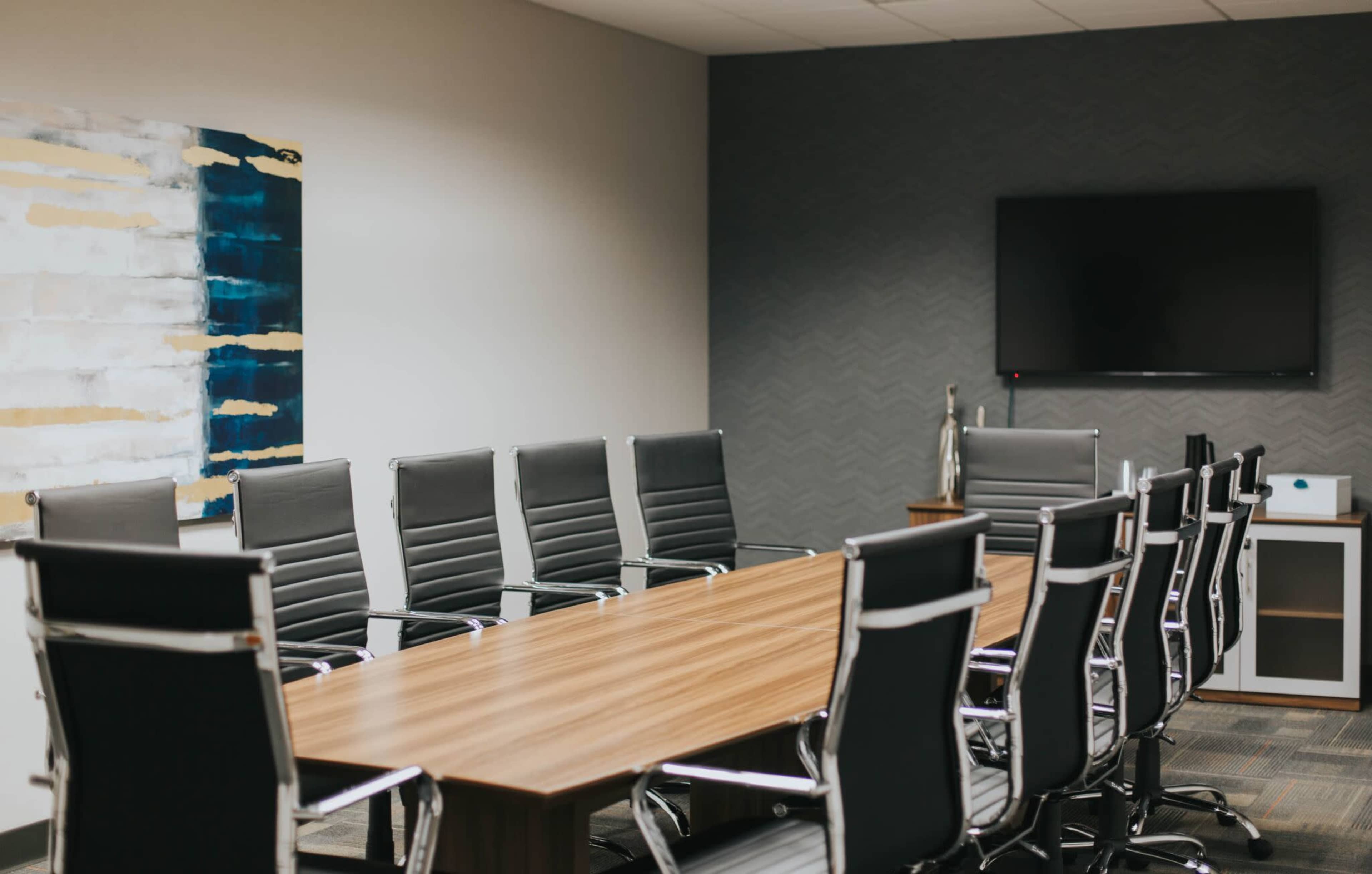A modern conference room features a wooden table surrounded by executive-style leather chairs and a wall-mounted television.
