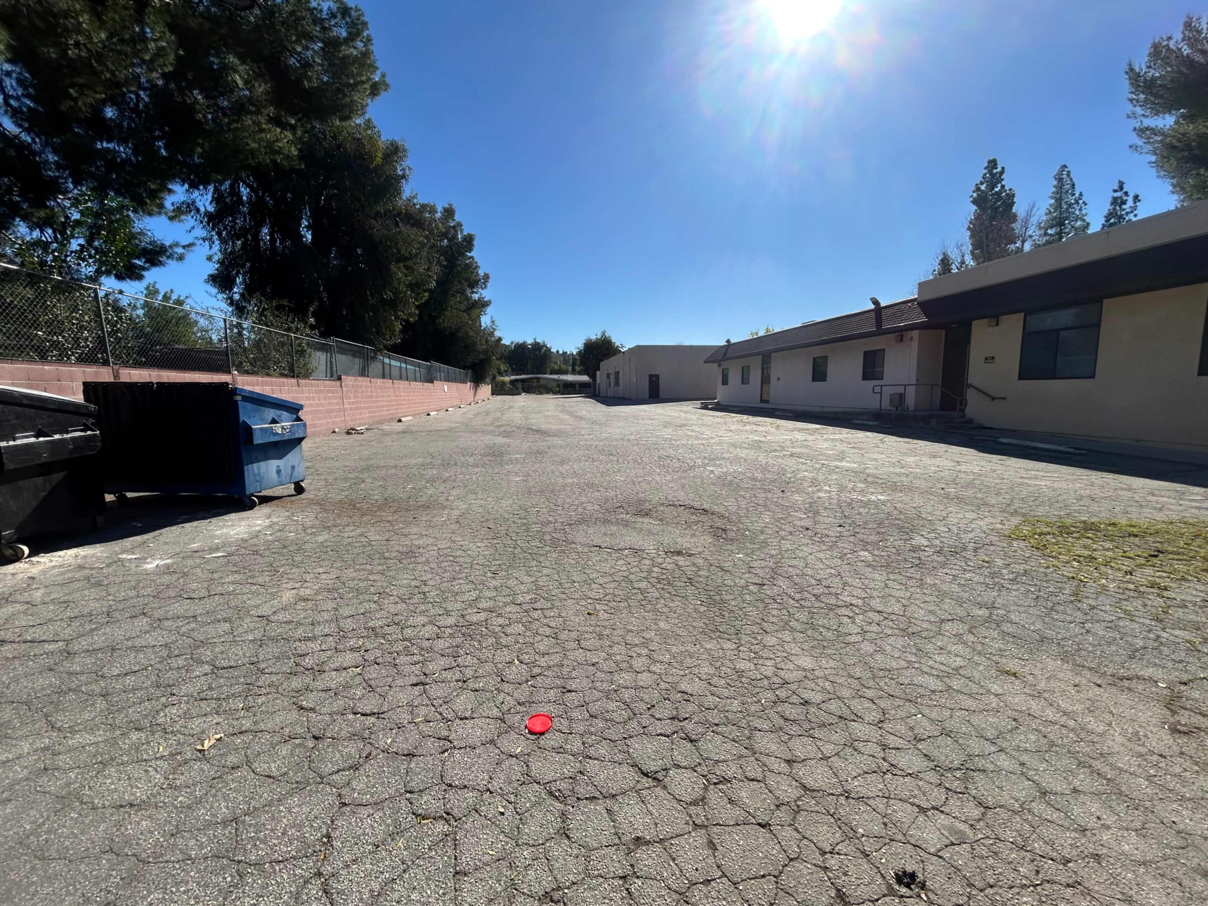 The image shows a vacant, cracked asphalt area with two buildings and several dumpsters on either side under clear blue skies.