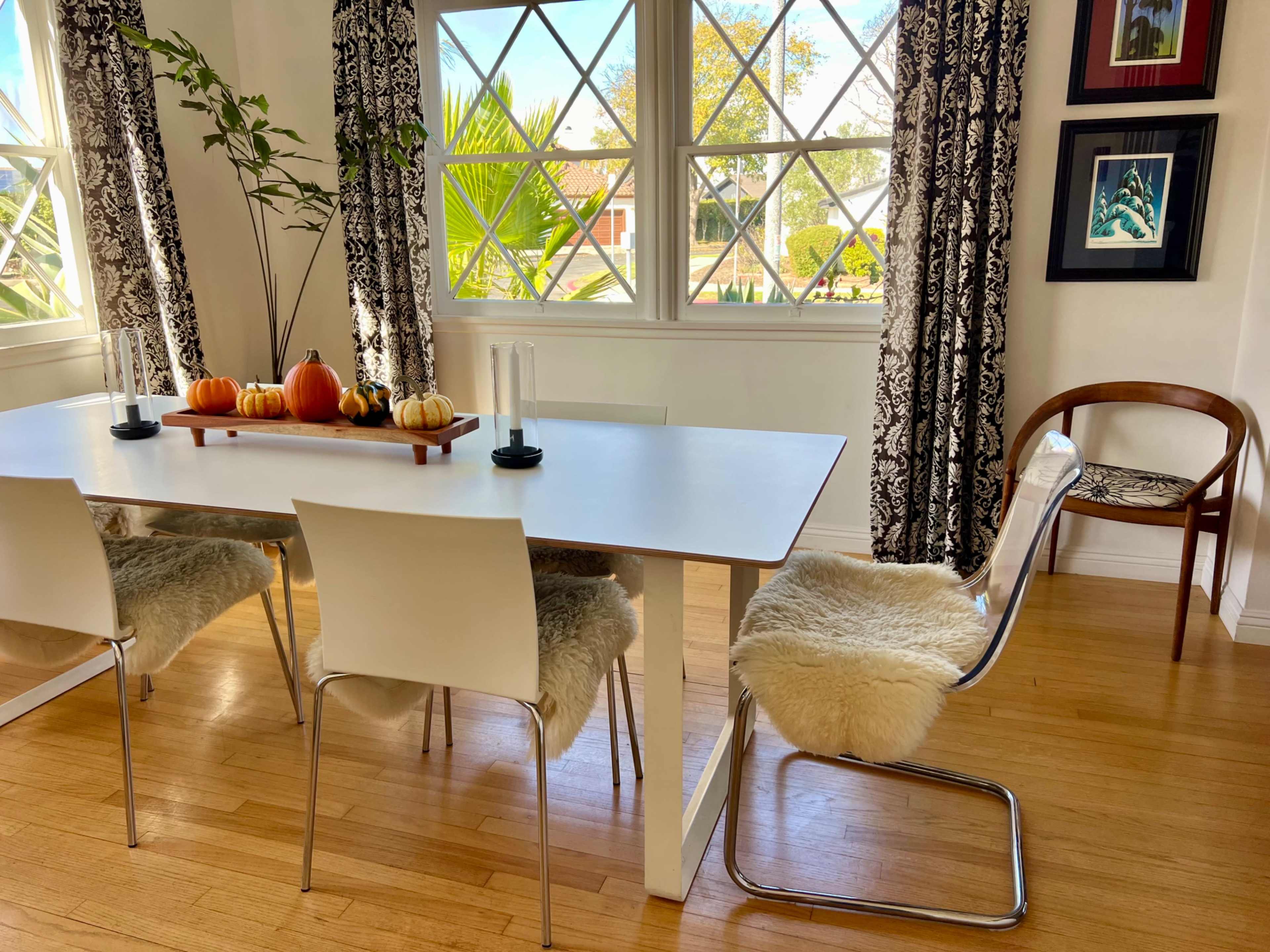 A dining room features a light-colored table with a decorative centerpiece of pumpkins and candles, surrounded by four chairs and large windows letting in natural light.