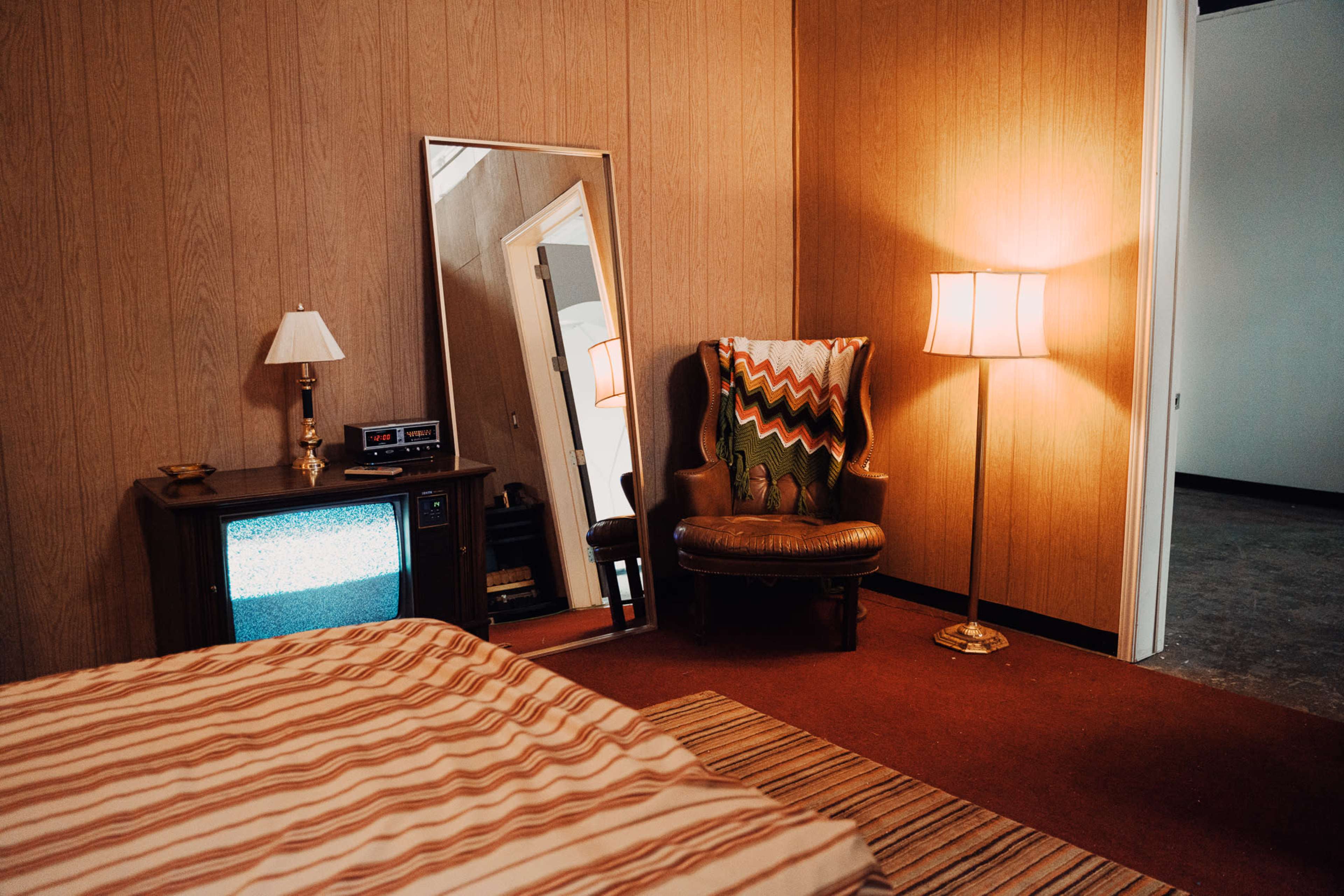 A vintage bedroom with a striped bedspread, a small television, a mirror, a lamp, and an upholstered chair with a patterned throw.