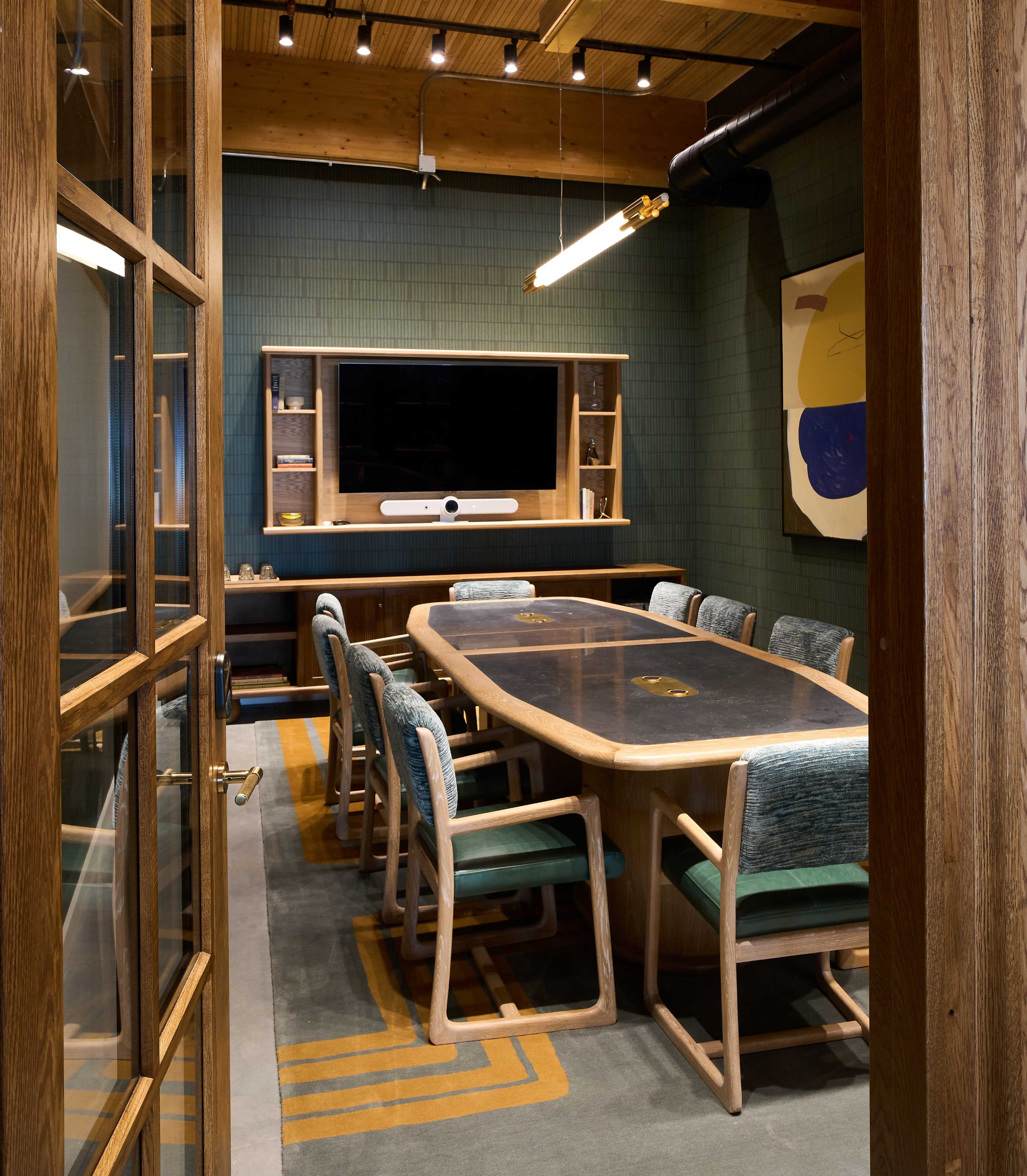 A modern conference room features a large wooden table surrounded by upholstered chairs, with a television mounted on the wall and a decorative carpet.