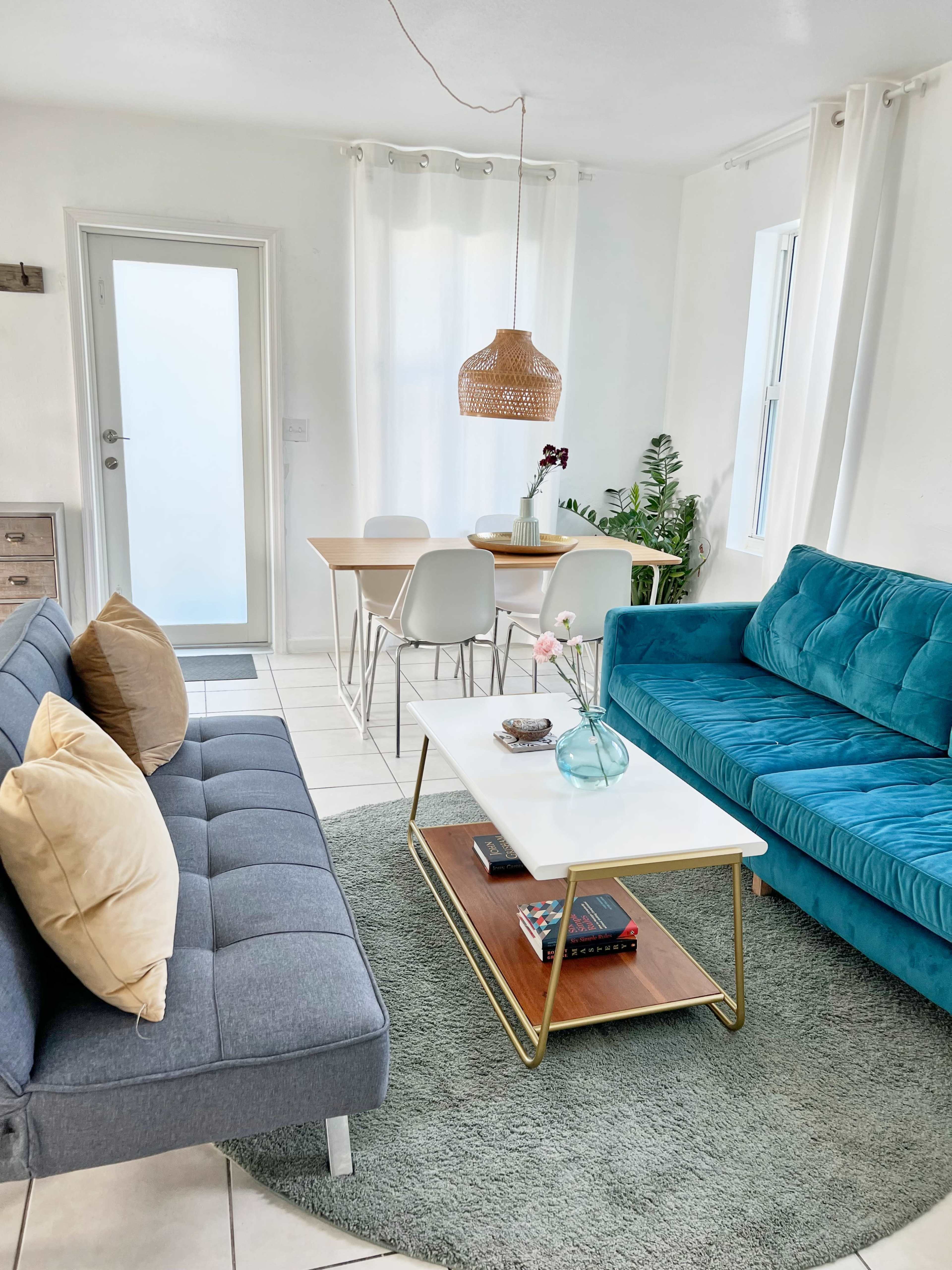The image shows a bright living room featuring a gray sofa with pillows, a teal couch, a glass coffee table on a round green rug, and a dining area with a wooden table and white chairs.