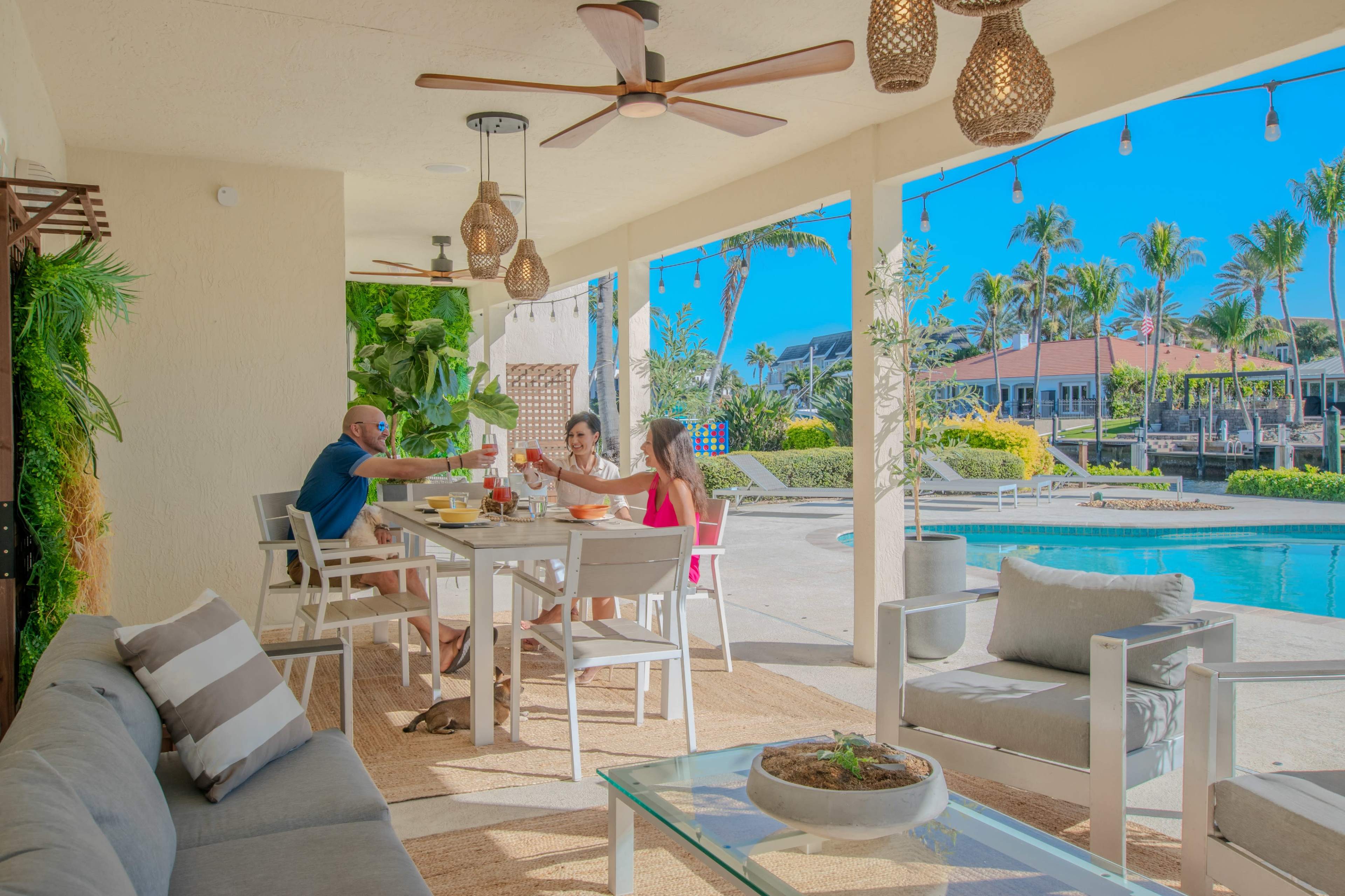 A group of three people enjoys a breakfast gathering on a patio by a swimming pool, surrounded by palm trees and a sunny blue sky.