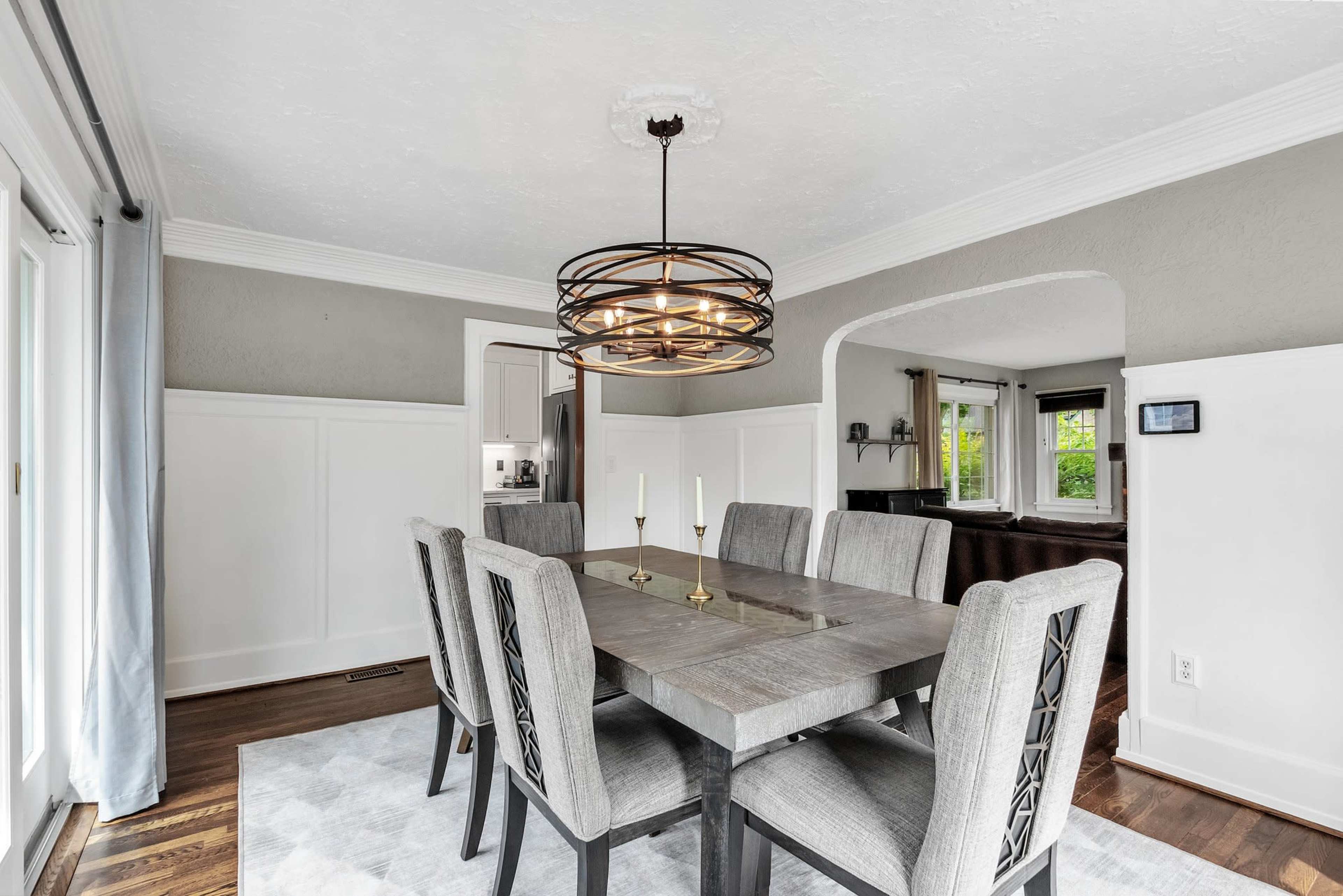 A modern dining room features a large table surrounded by six chairs, with a geometric light fixture hanging above and large windows letting in natural light.