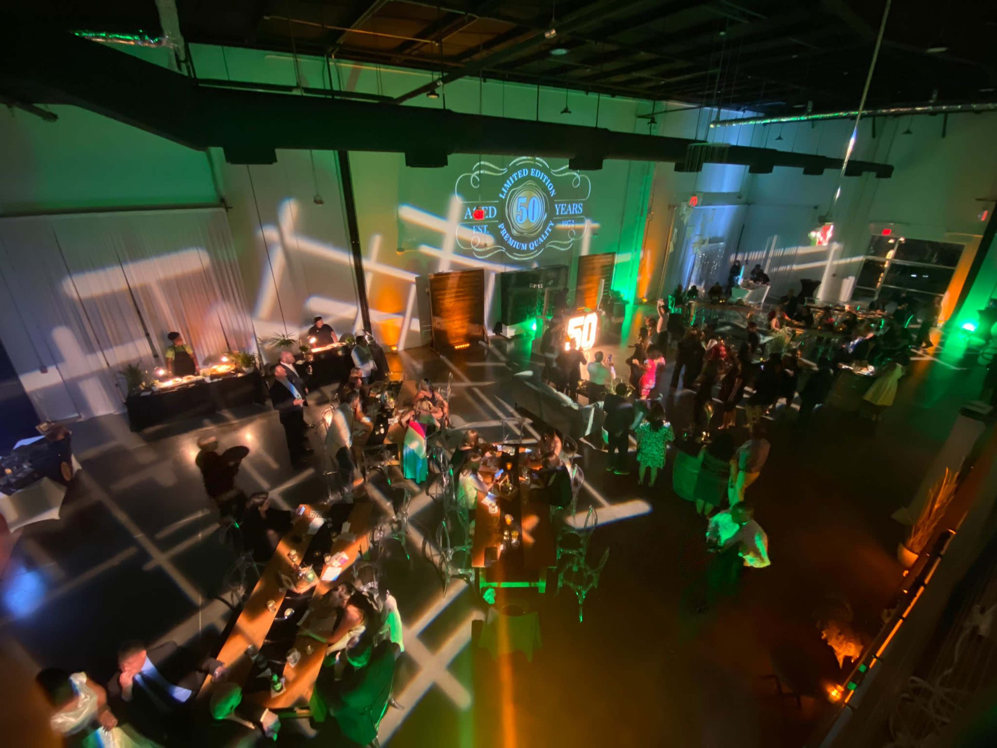 The image shows a lively indoor event space filled with guests celebrating a milestone, featuring tables, a buffet area, and a large projection displaying "60 Years."