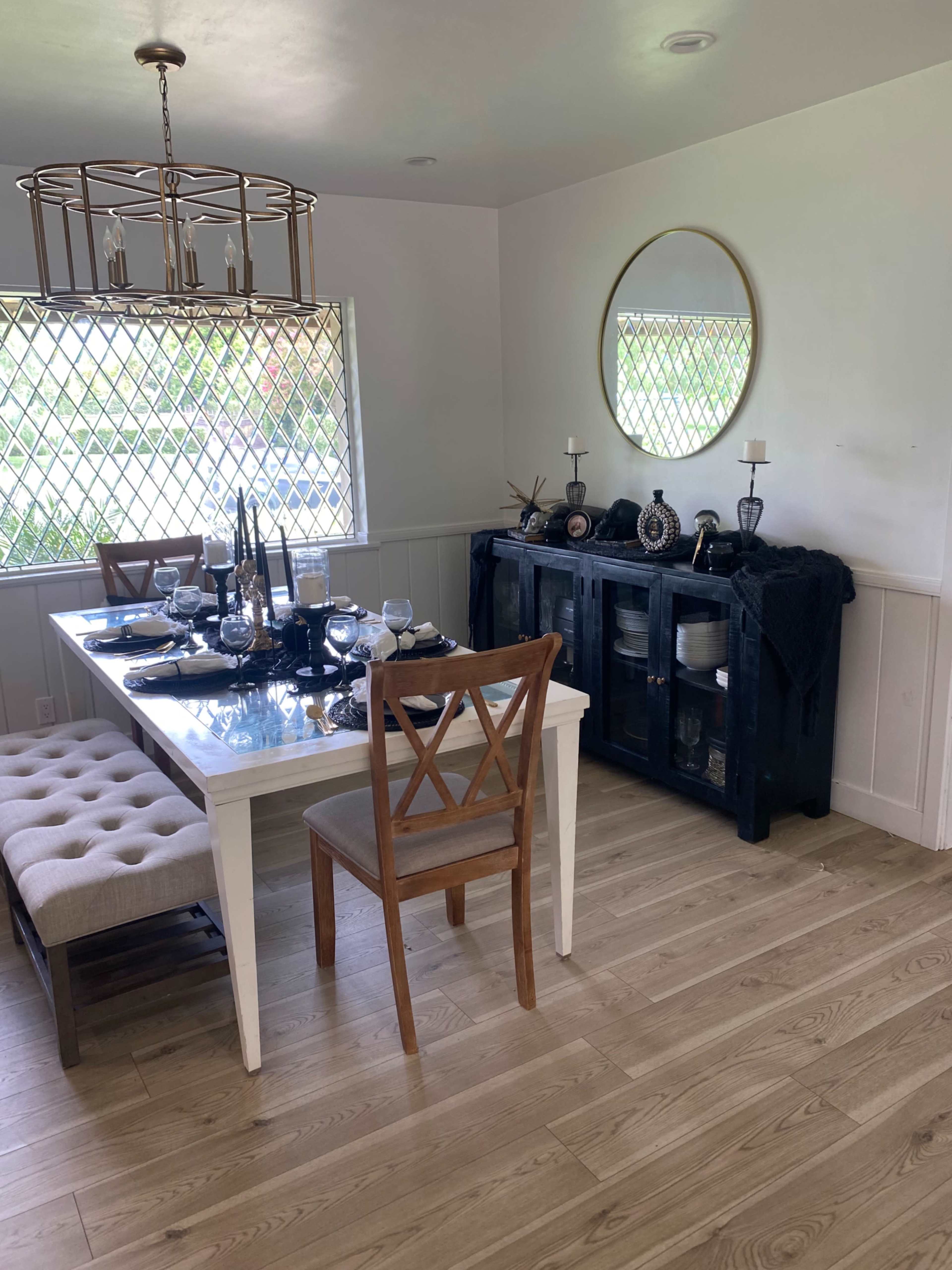 A dining room features a white table set for a meal, wooden chairs, and a black sideboard, with a large mirror and a chandelier overhead.