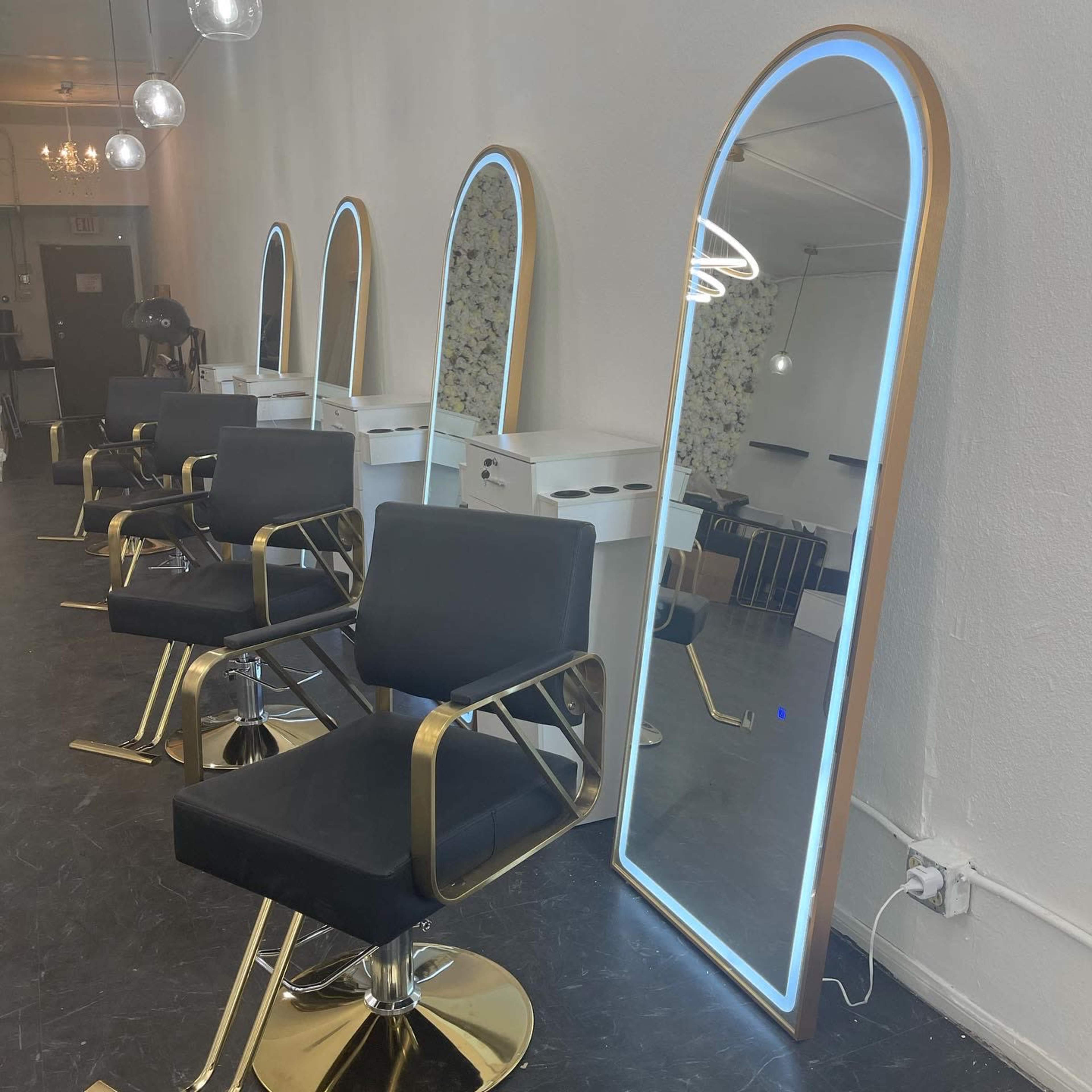 A modern hair salon interior featuring several styling chairs and a large illuminated mirror along the wall.