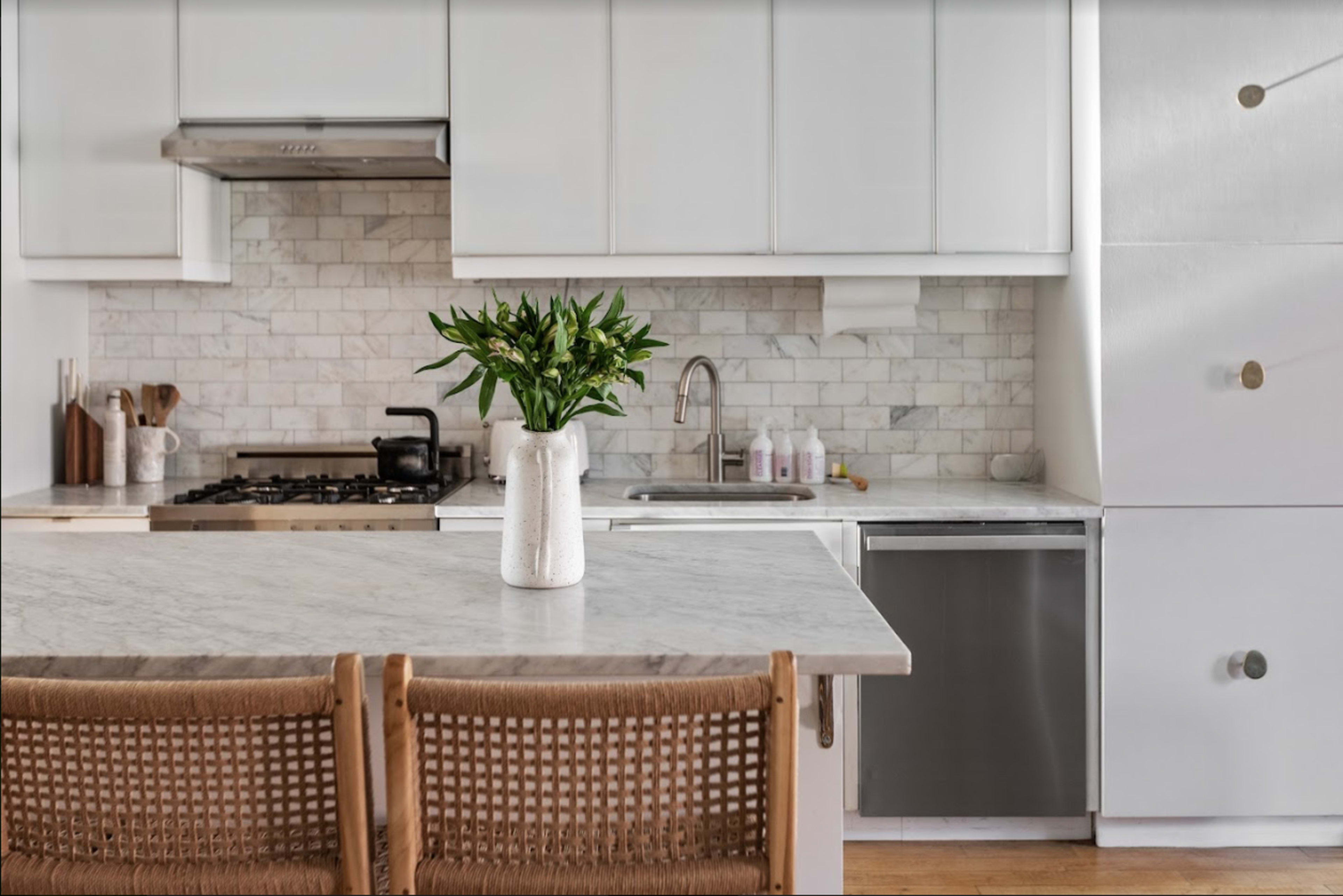A clean, modern kitchen features a marble countertop, white cabinetry, and a vase of greenery on the island.