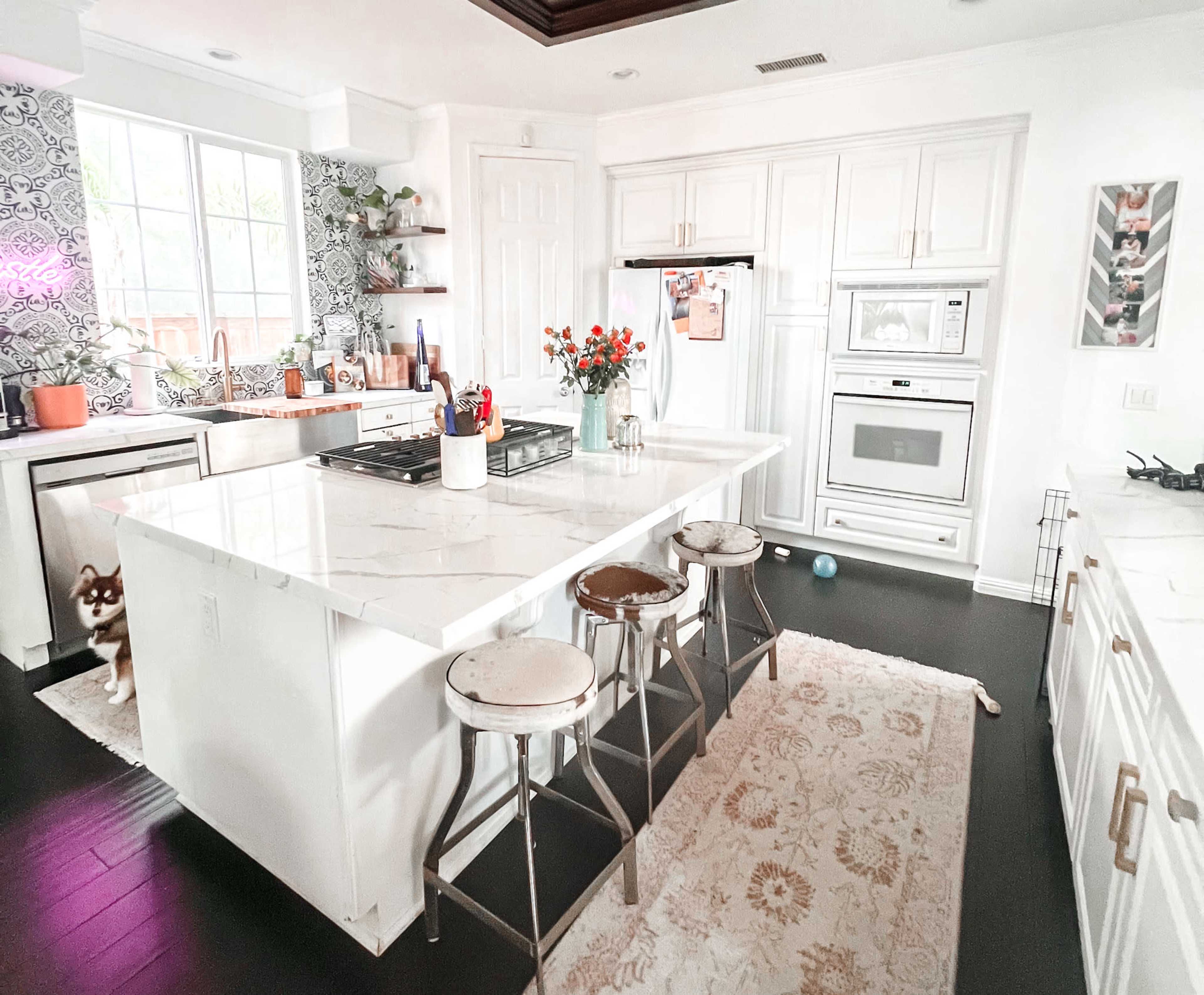 The image shows a modern kitchen with a large white island, three metal stools, and a variety of appliances and plants in the surrounding area.