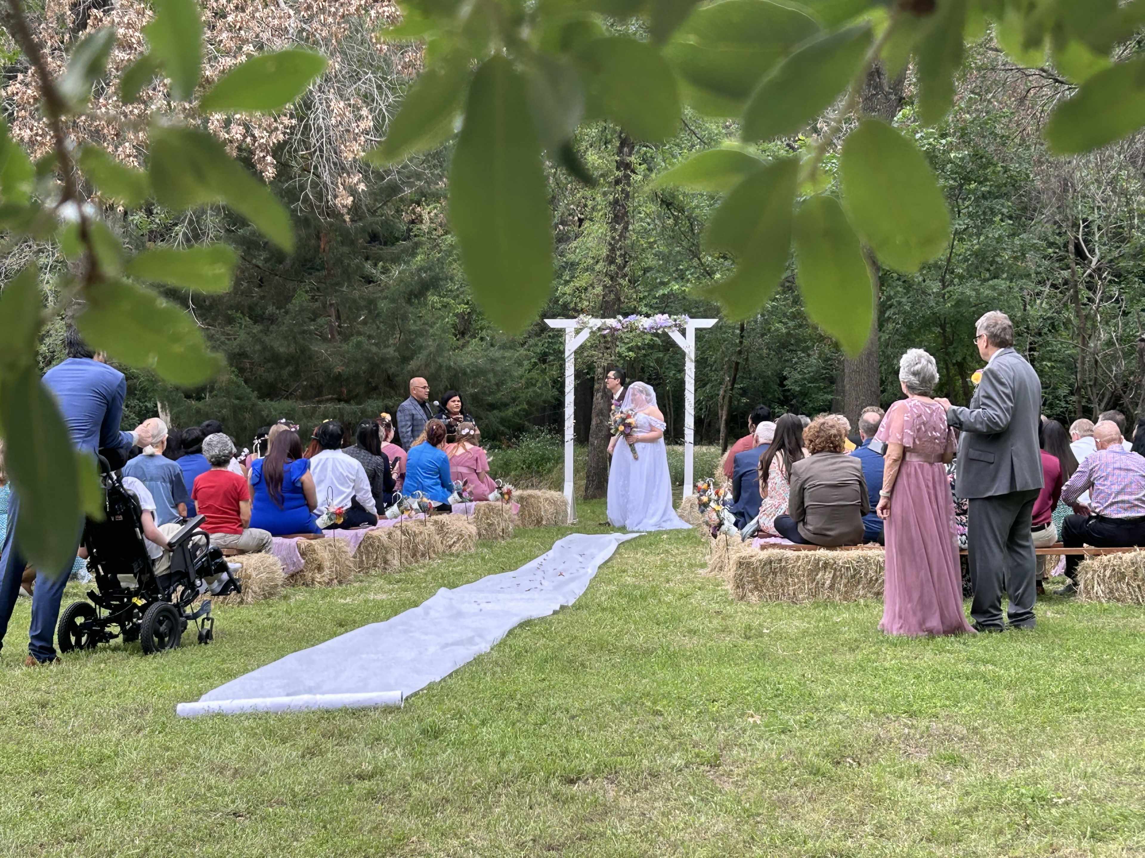 A bride walks down a white path towards an altar in a grassy outdoor setting, where guests are seated on straw bales.