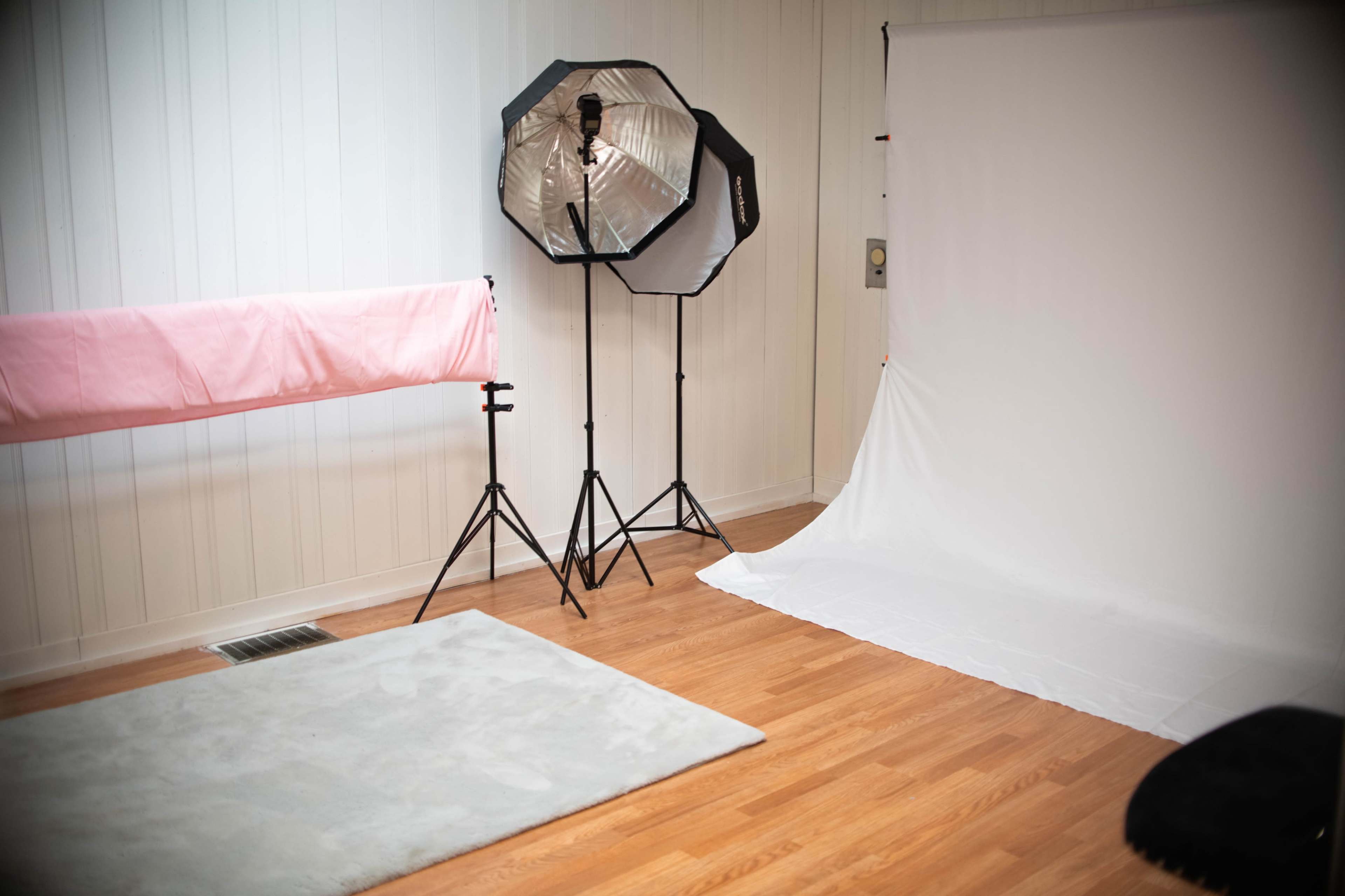 The image shows a photography studio with a pink backdrop, two softbox lights, and a white backdrop on the floor.