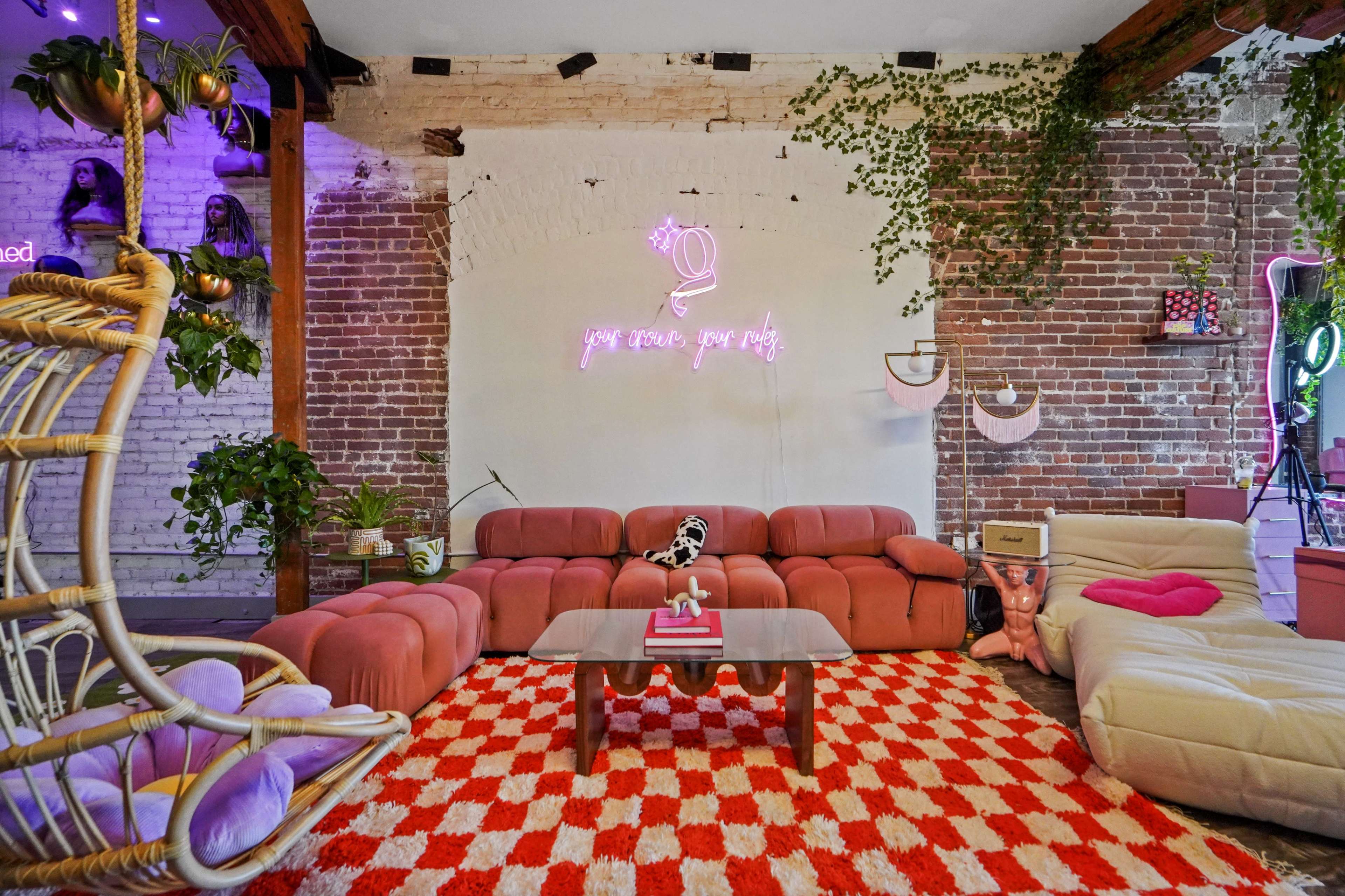 A cozy lounge area with a pink sectional sofa, a checkered rug, hanging plants, and neon wall art against a brick backdrop.