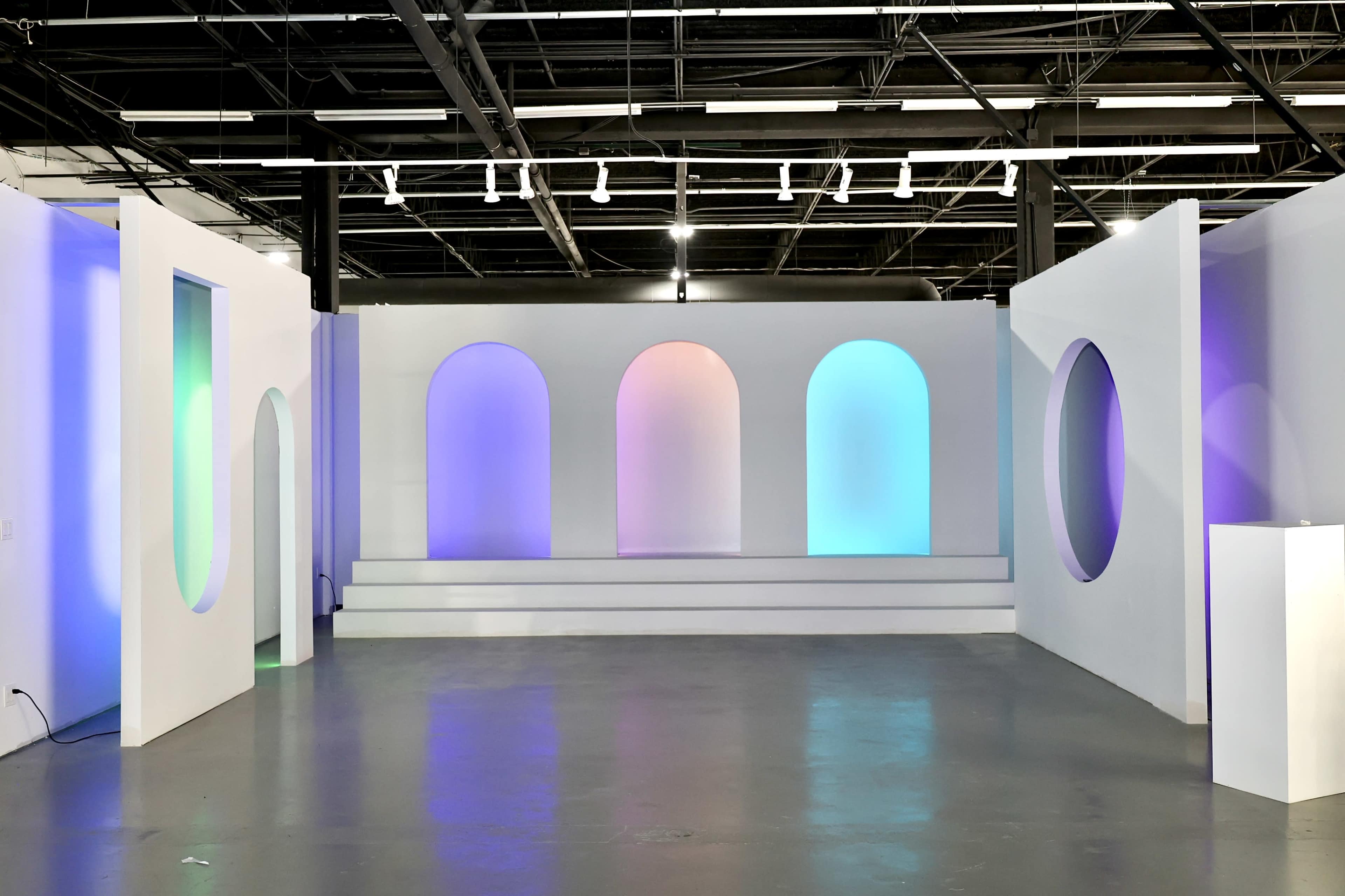 The image shows a spacious gallery with white walls featuring arched cutouts, illuminated by soft, colored lighting.
