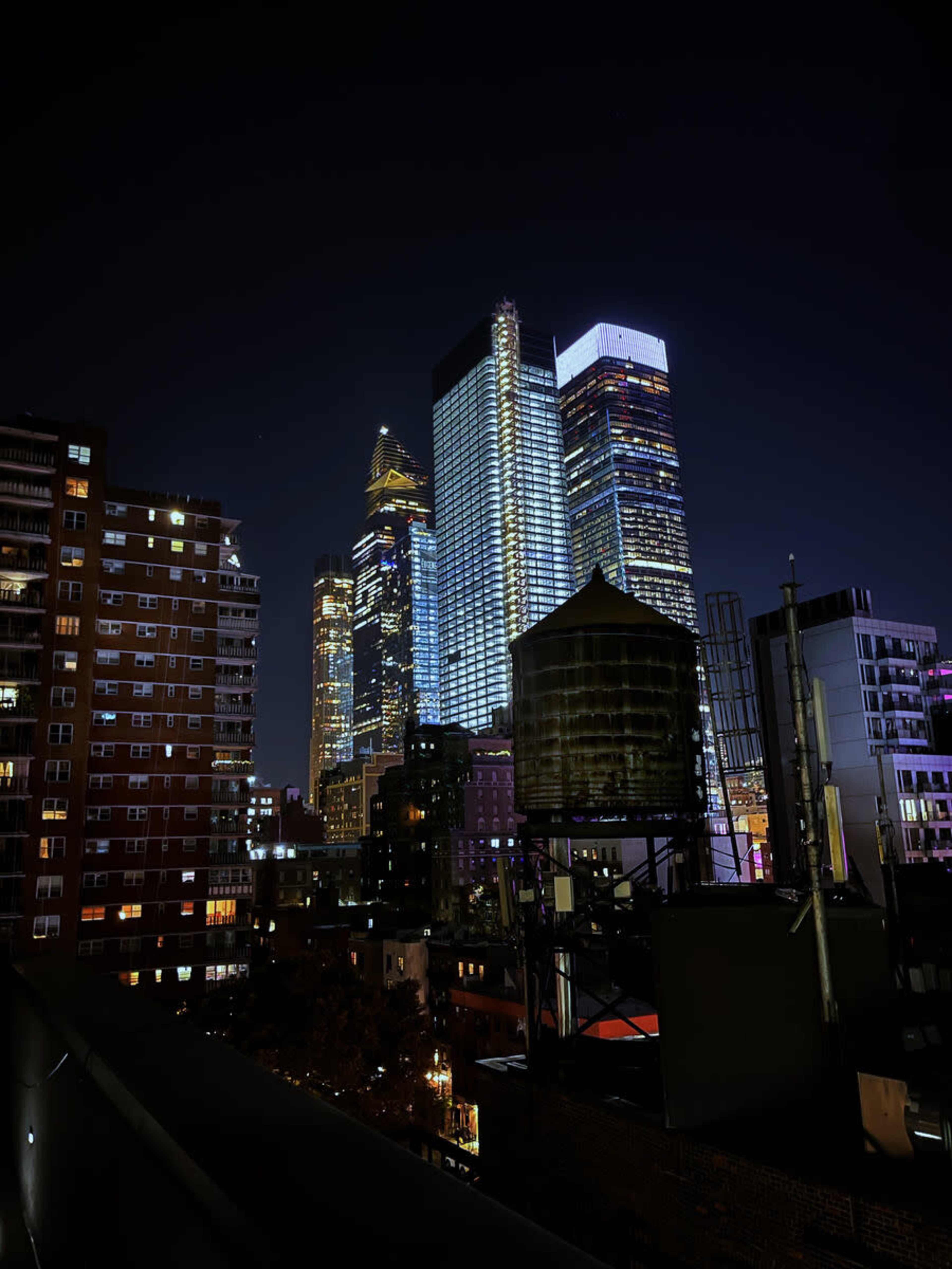 A city skyline at night, featuring tall illuminated skyscrapers alongside industrial rooftops and water towers.