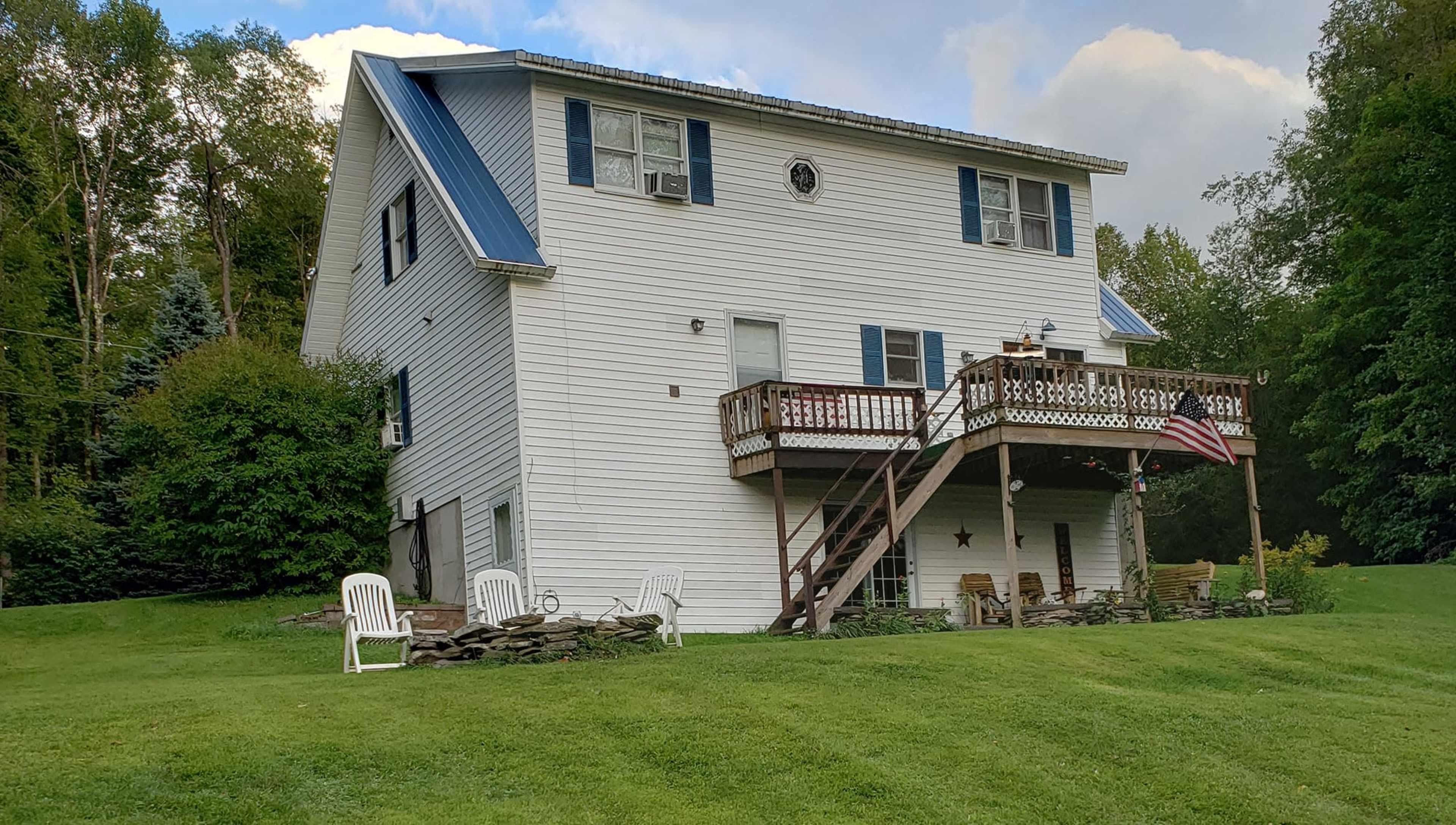 A two-story white house with a blue metal roof features a front deck and an American flag, surrounded by a grassy lawn and several white chairs.