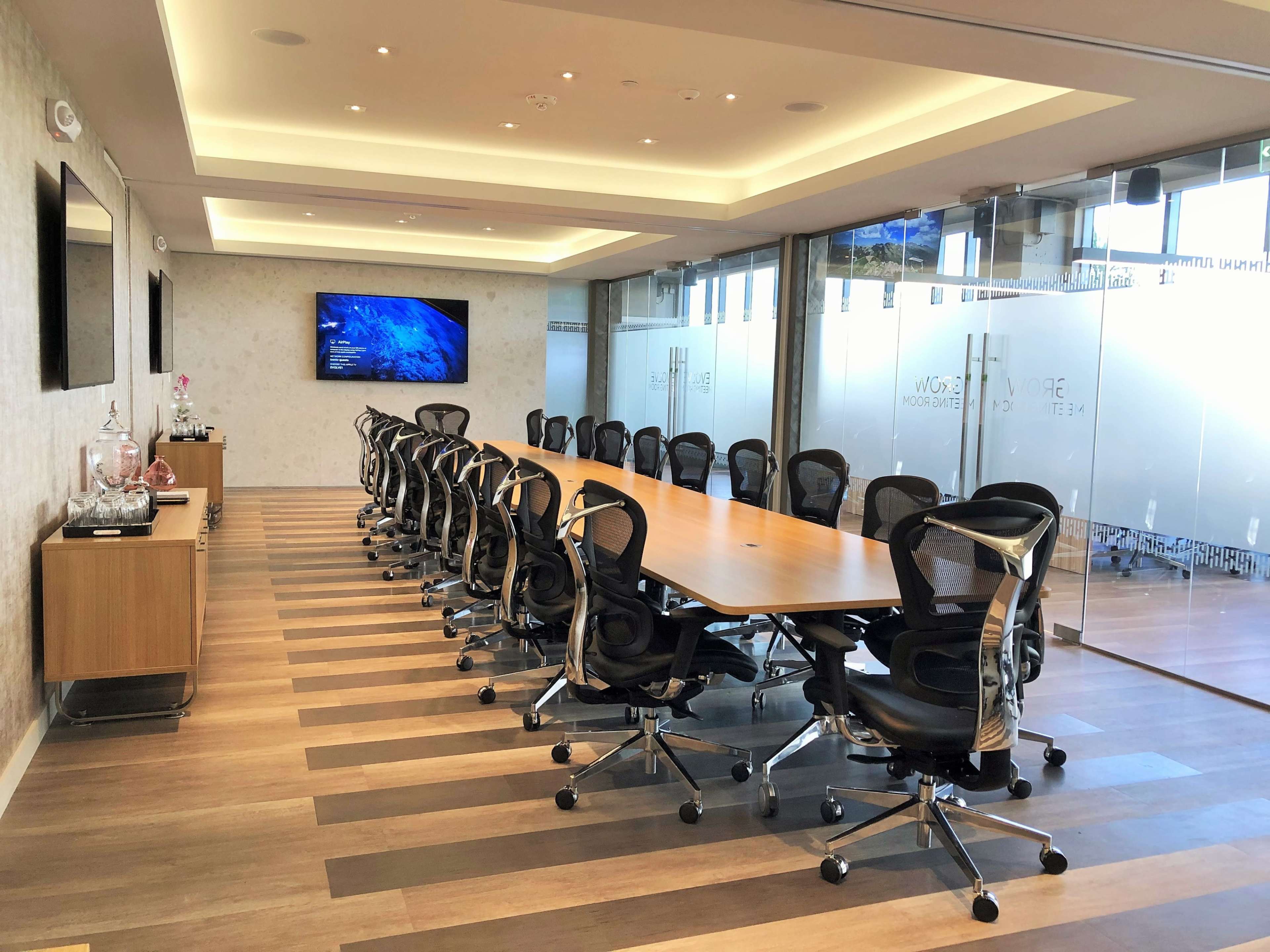 A large conference room features a long wooden table surrounded by ergonomic chairs and equipped with a wall-mounted screen.
