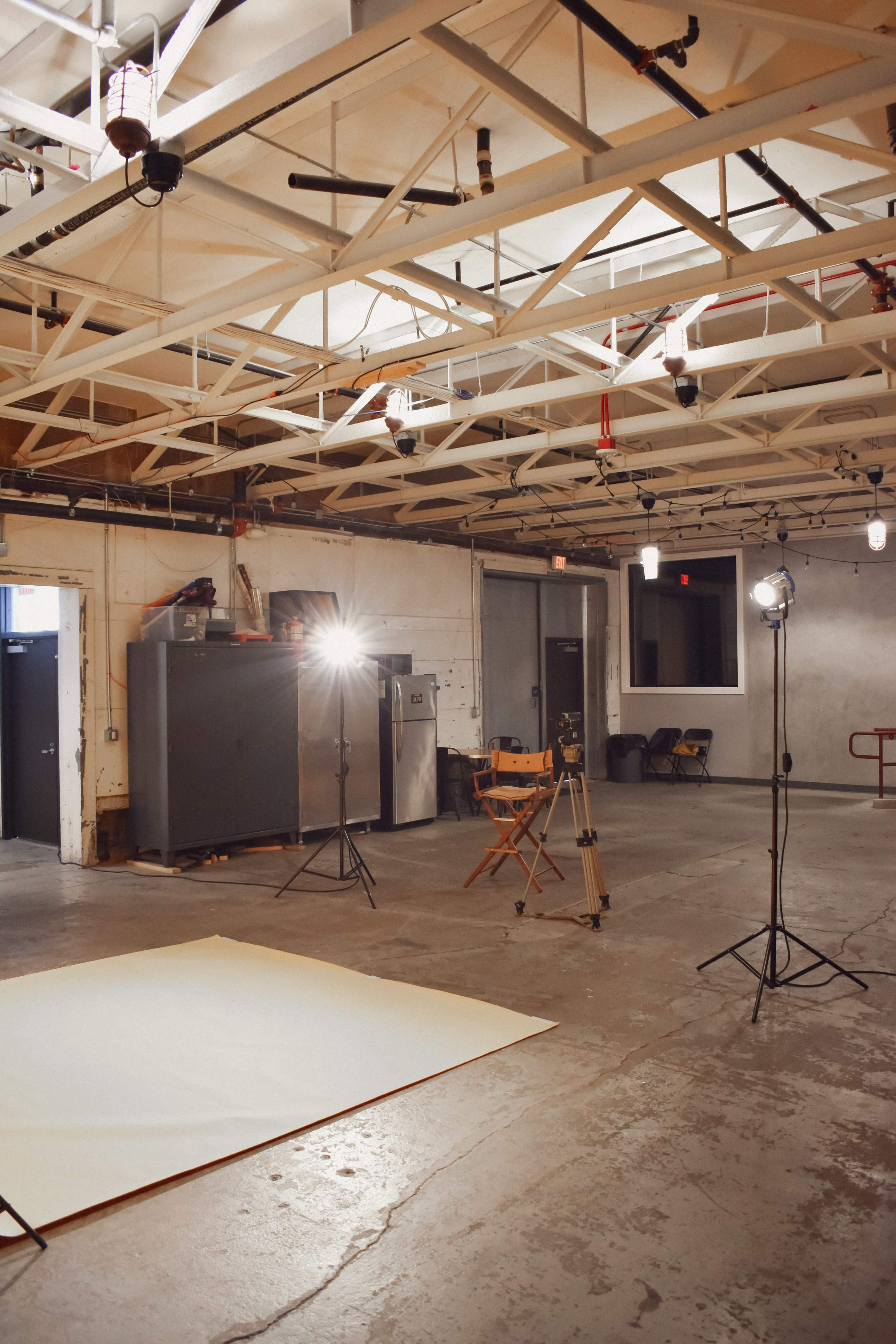 A spacious room features a backdrop on the floor, two lighting fixtures on stands, a director's chair, and various appliances against the walls.