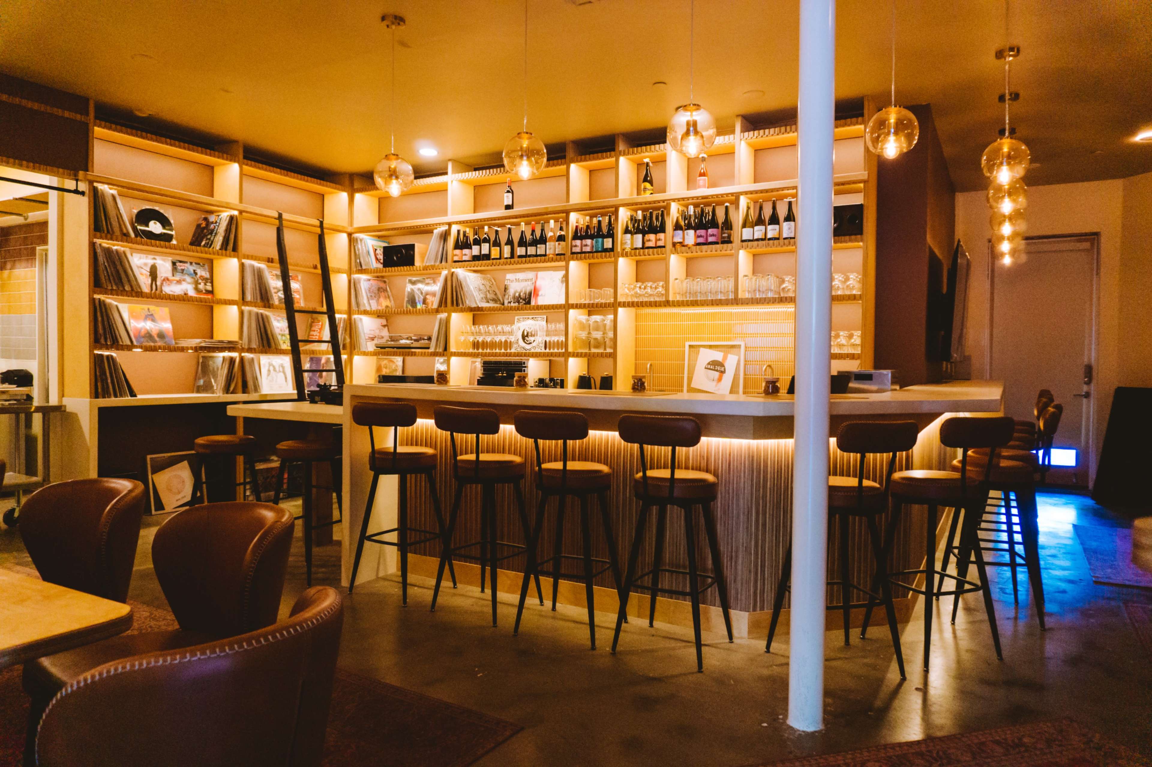A modern bar features a well-lit counter with high stools and shelves displaying bottles and vinyl records.