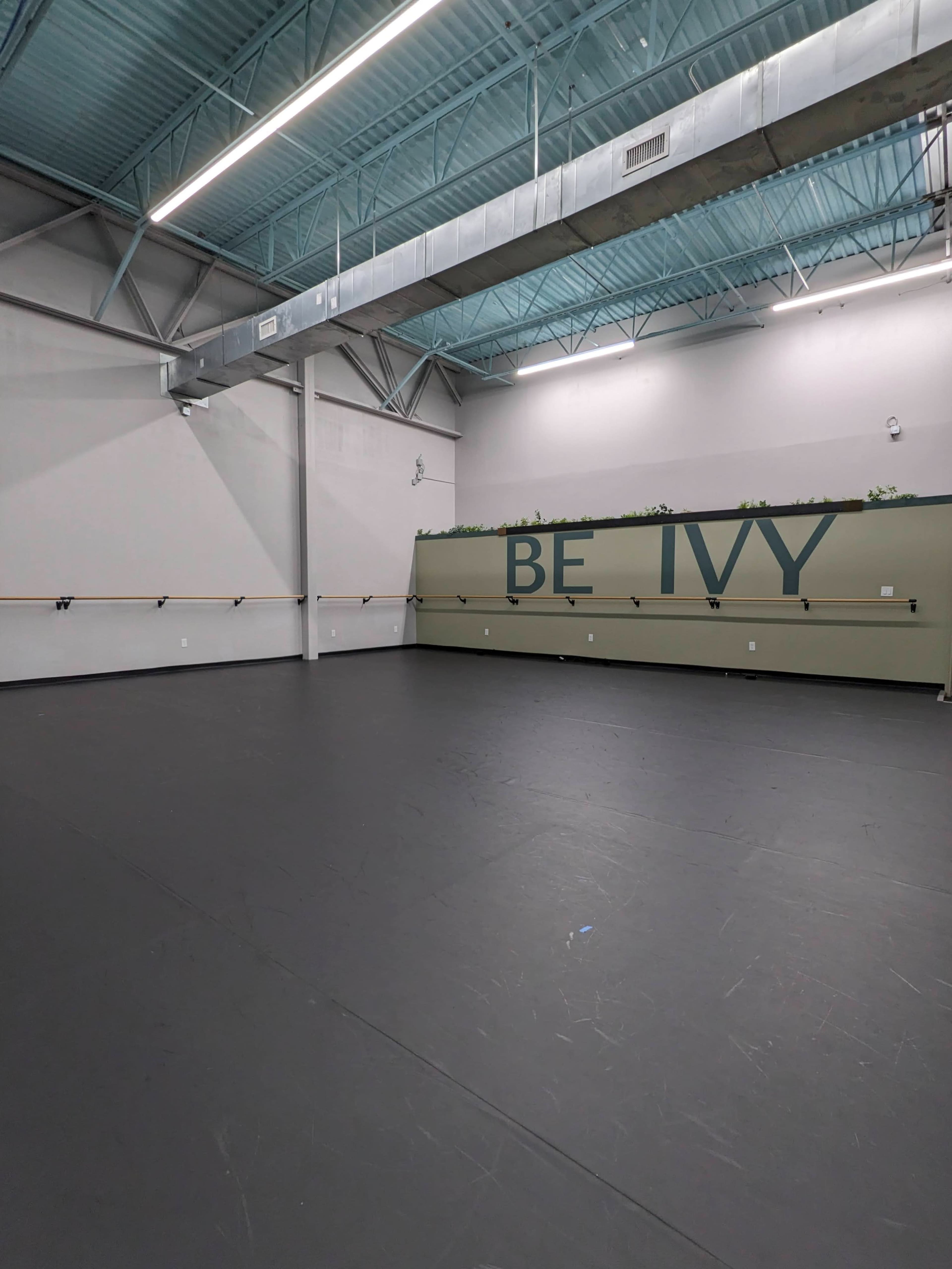 The image shows an empty dance studio with a black floor, mirrored walls, and the words "BE IVY" displayed on one wall.