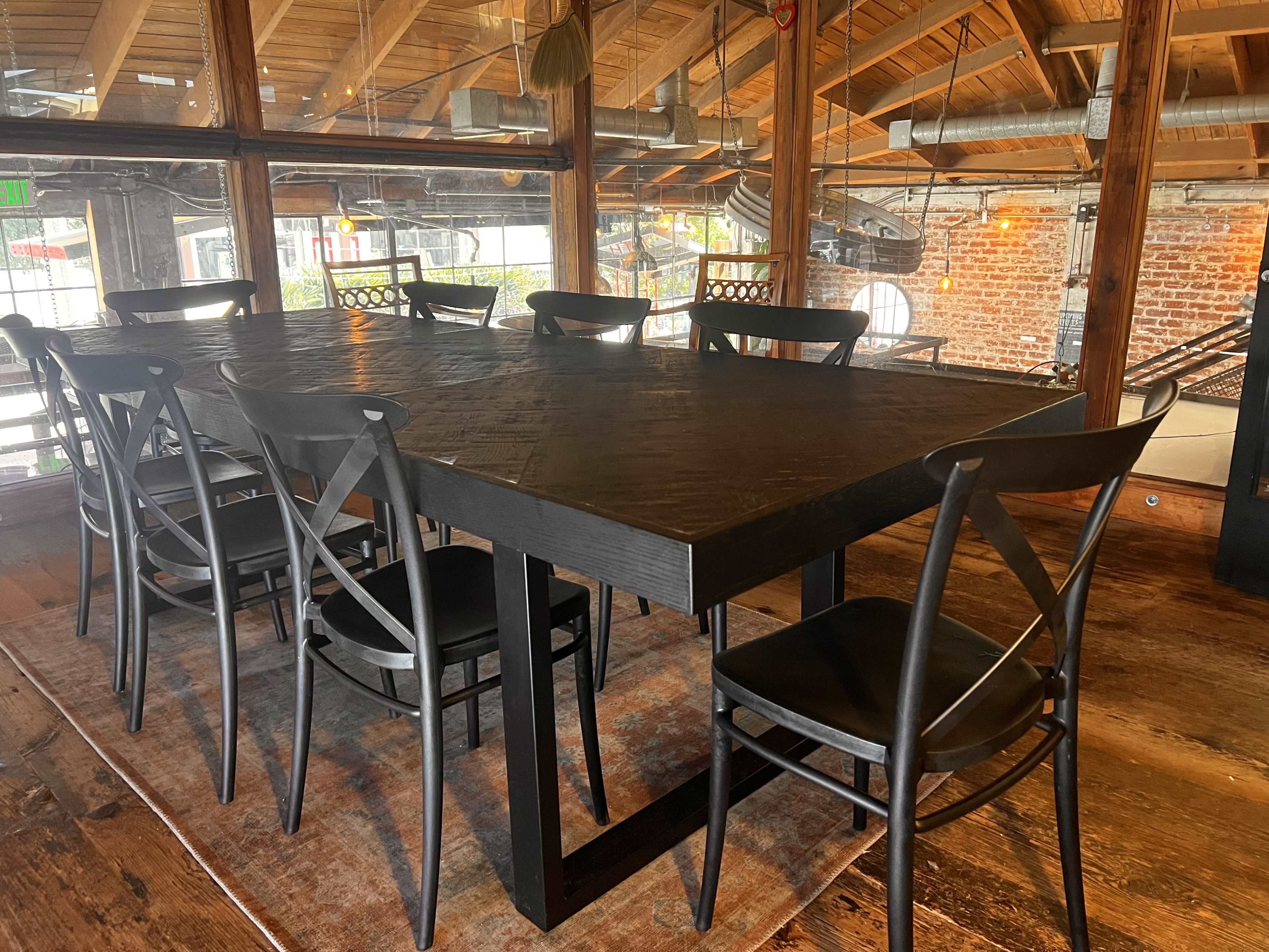 A large black wooden dining table surrounded by several black chairs is set in a rustic restaurant with exposed brick and wooden beams.