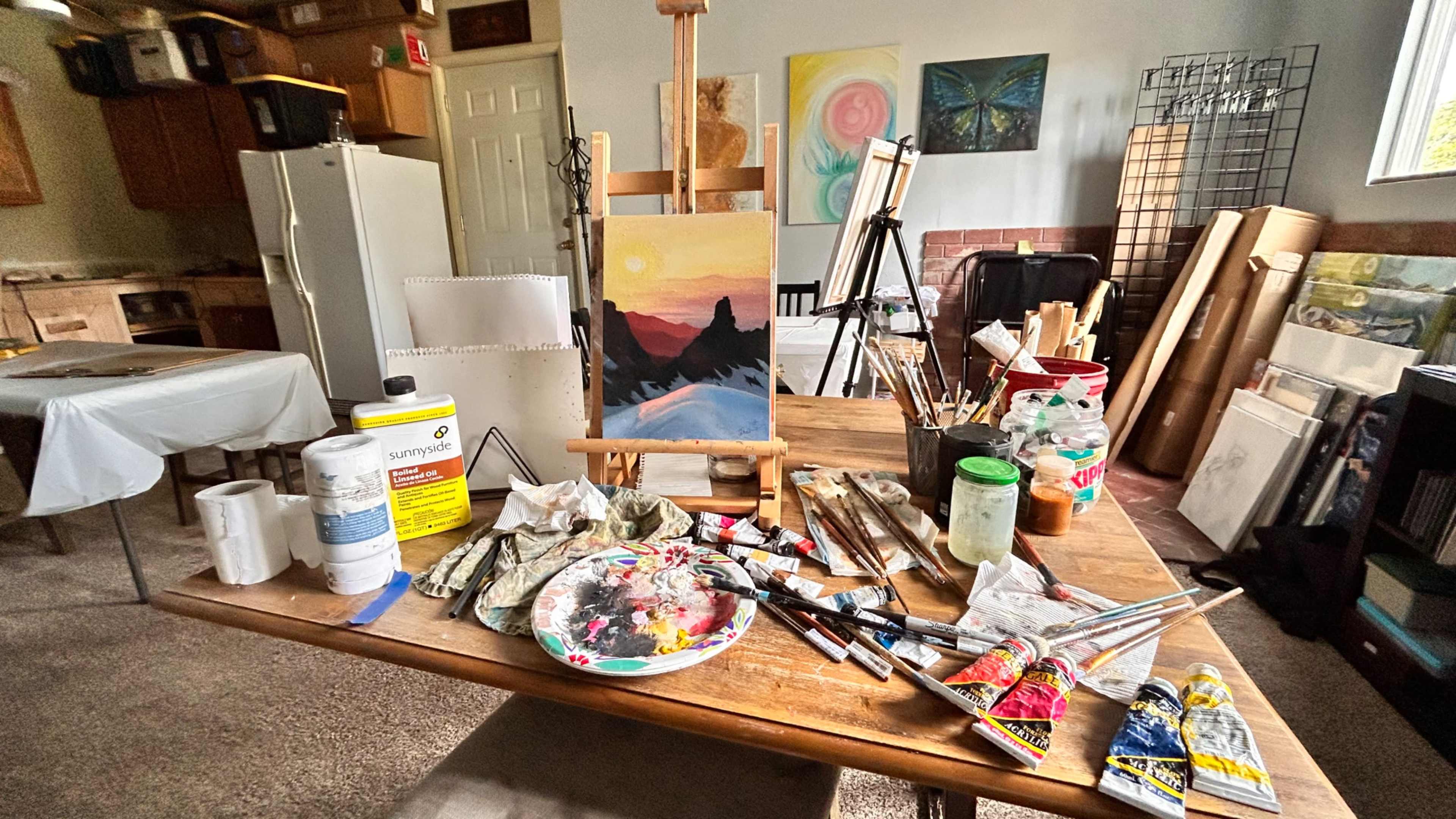 An artist's workspace is filled with paint tubes, brushes, a palette, and various unfinished canvases in a bright room with a kitchen in the background.