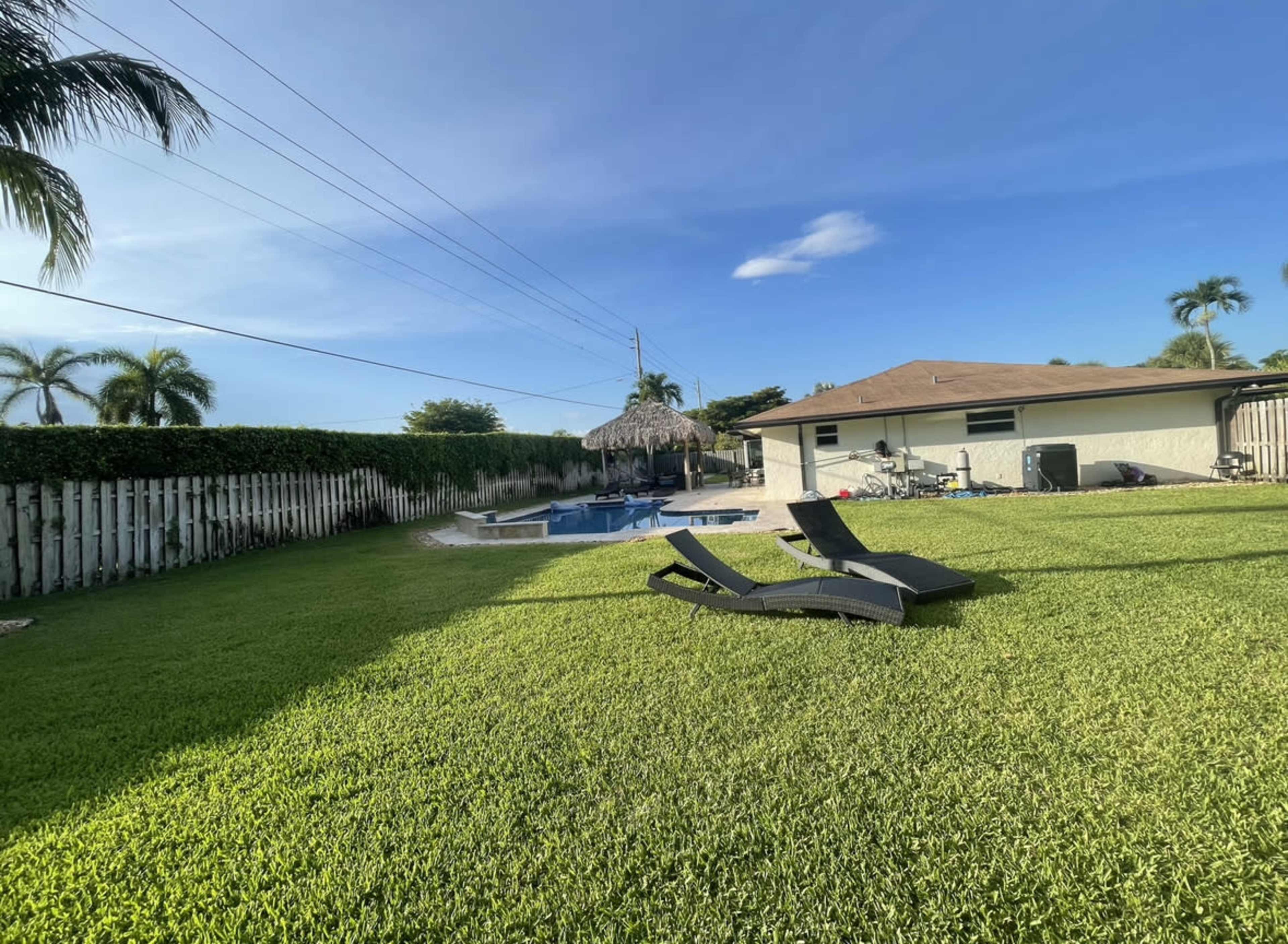A backyard features a pool surrounded by lounge chairs and a thatched-roof structure, with a neatly maintained lawn and a house in the background.