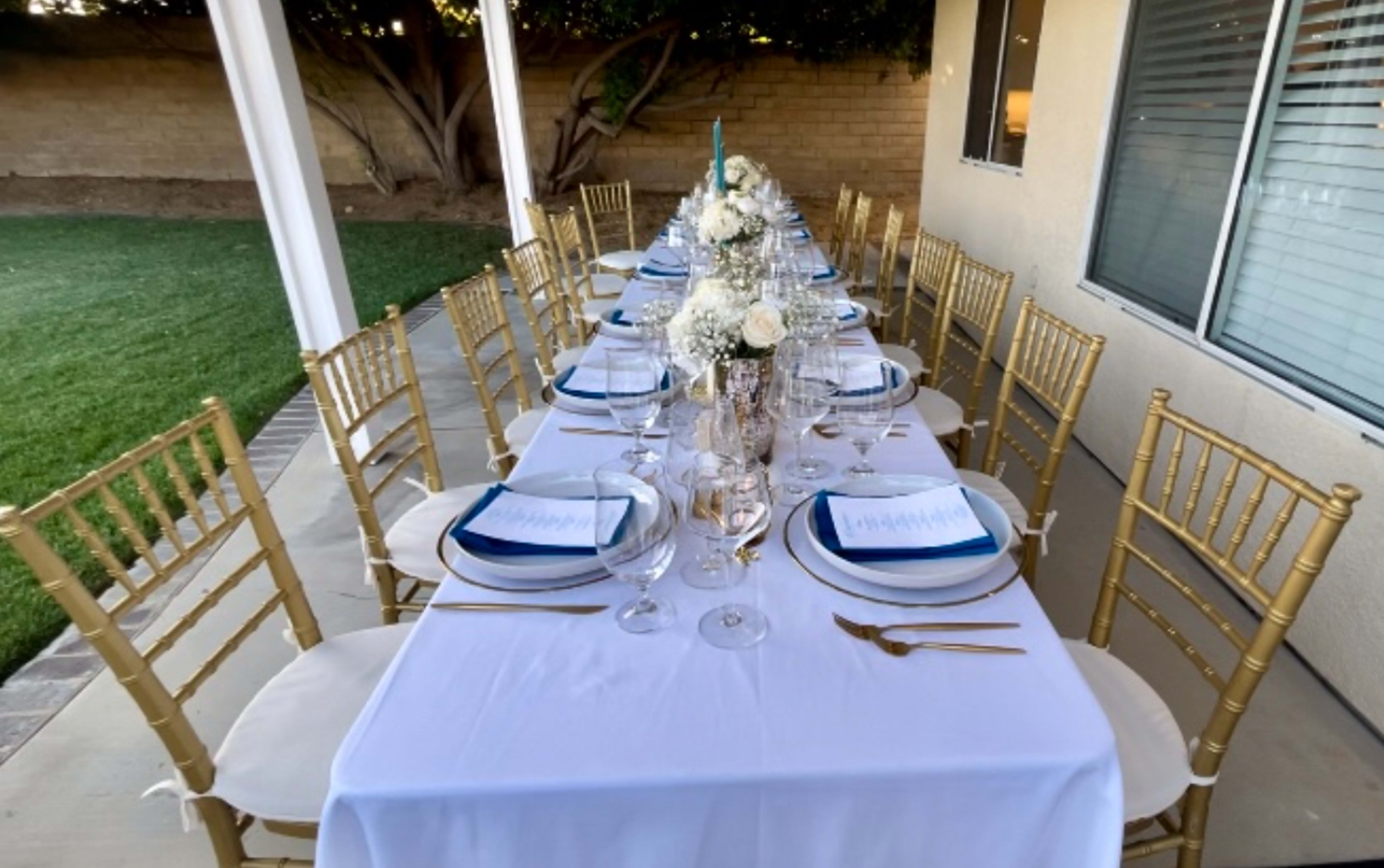 A long, elegantly set dining table with gold chairs, white tablecloth, and decorative centerpieces is arranged on a patio beside a grassy area.