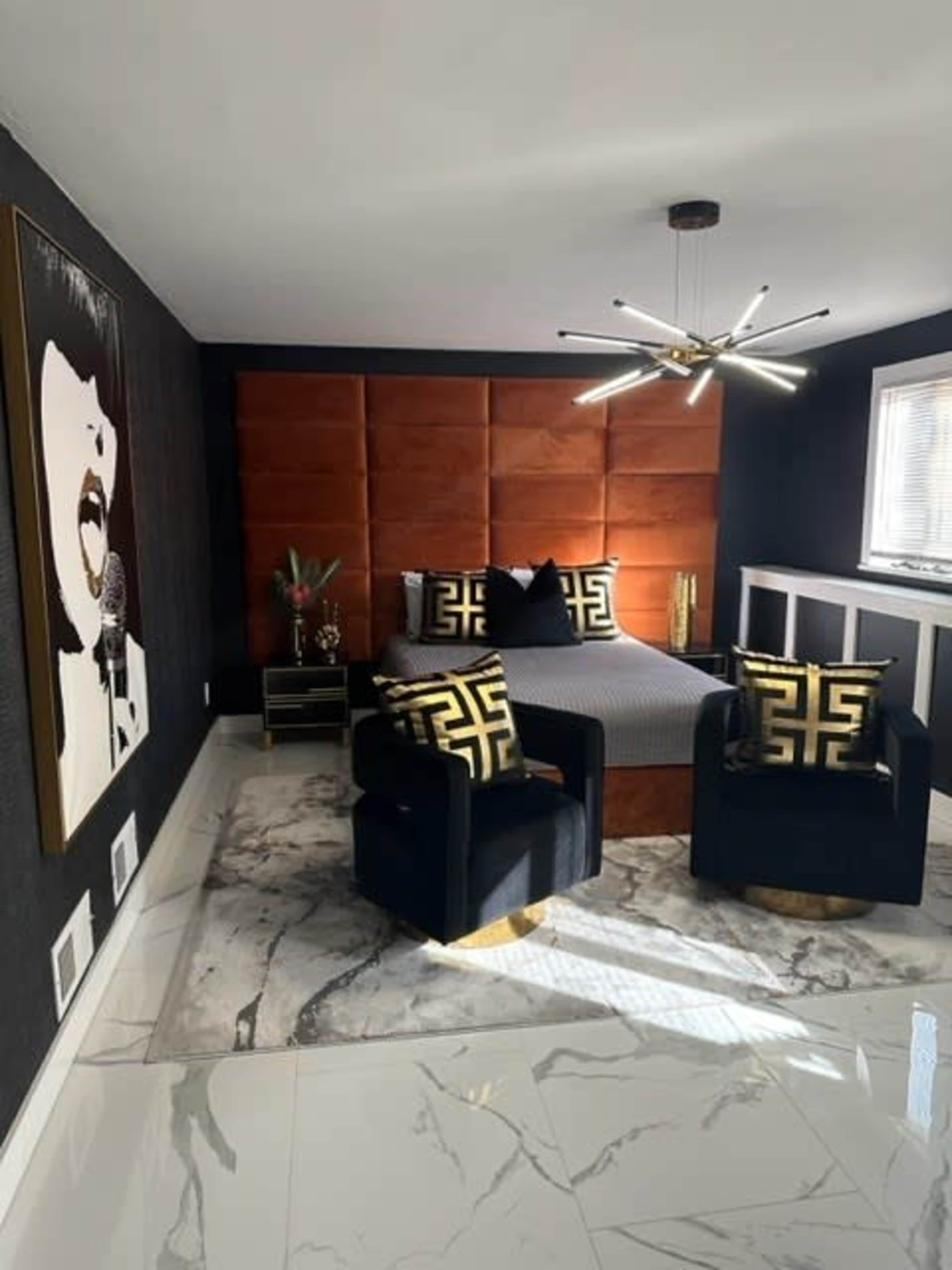 A modern bedroom with an orange upholstered headboard, a patterned bedspread, two black chairs with gold accents, and a large artwork on the wall.