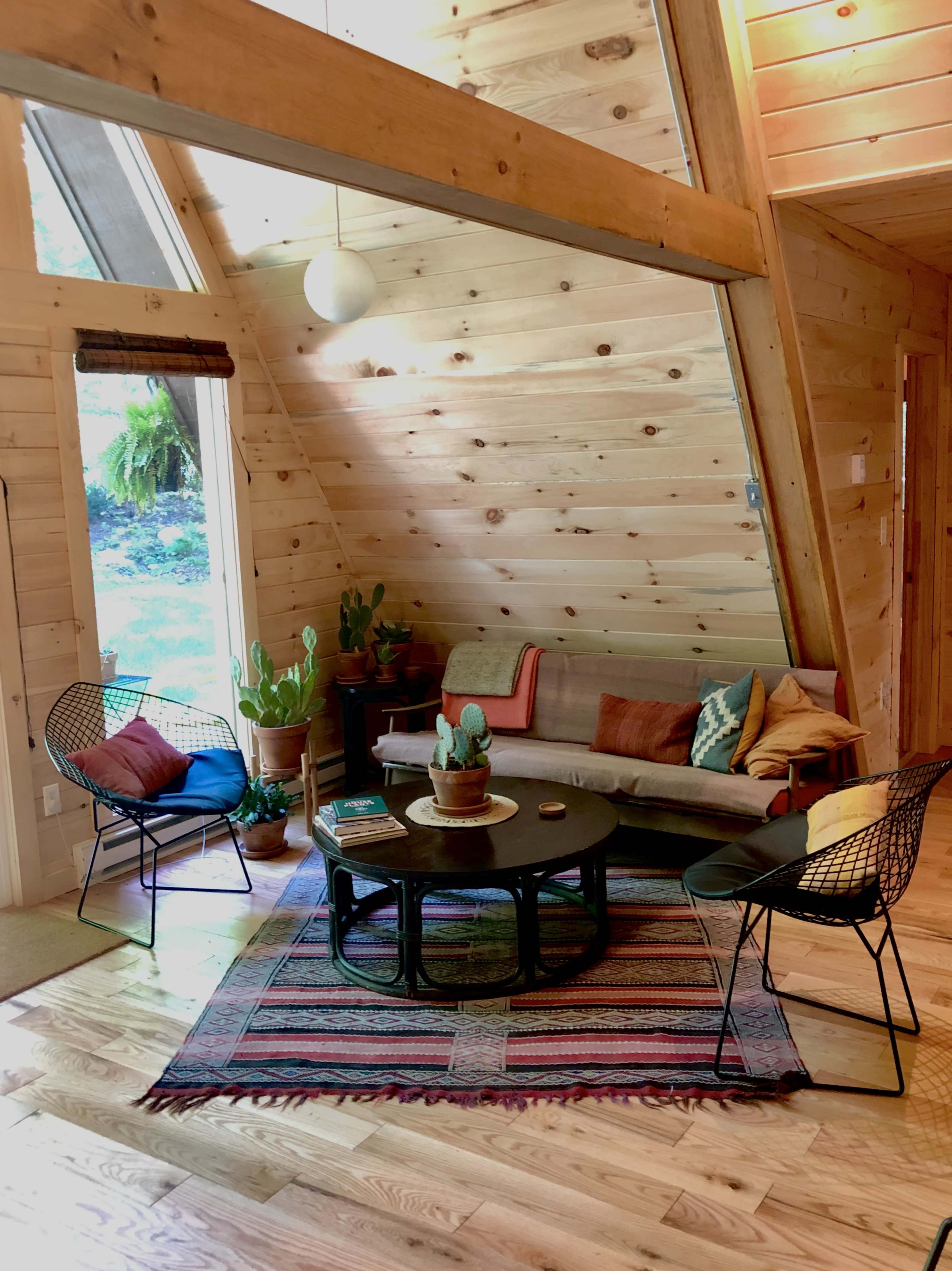 The interior features a wooden A-frame living room with a sofa, two wire chairs, a circular coffee table, and various potted plants, all arranged on a patterned area rug.